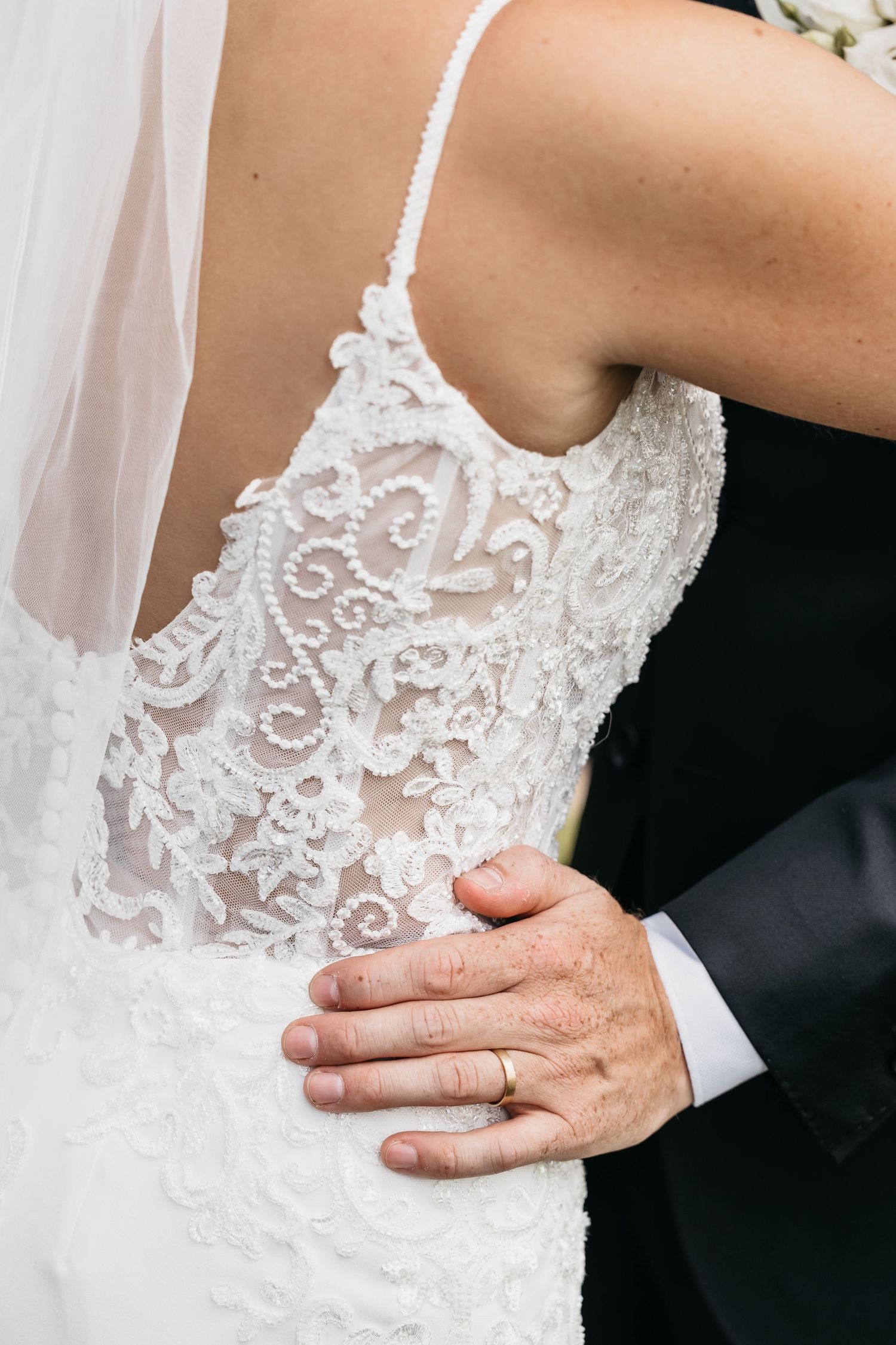 Groom placing hand on bride's lace wedding dress back with wedding ring visible.