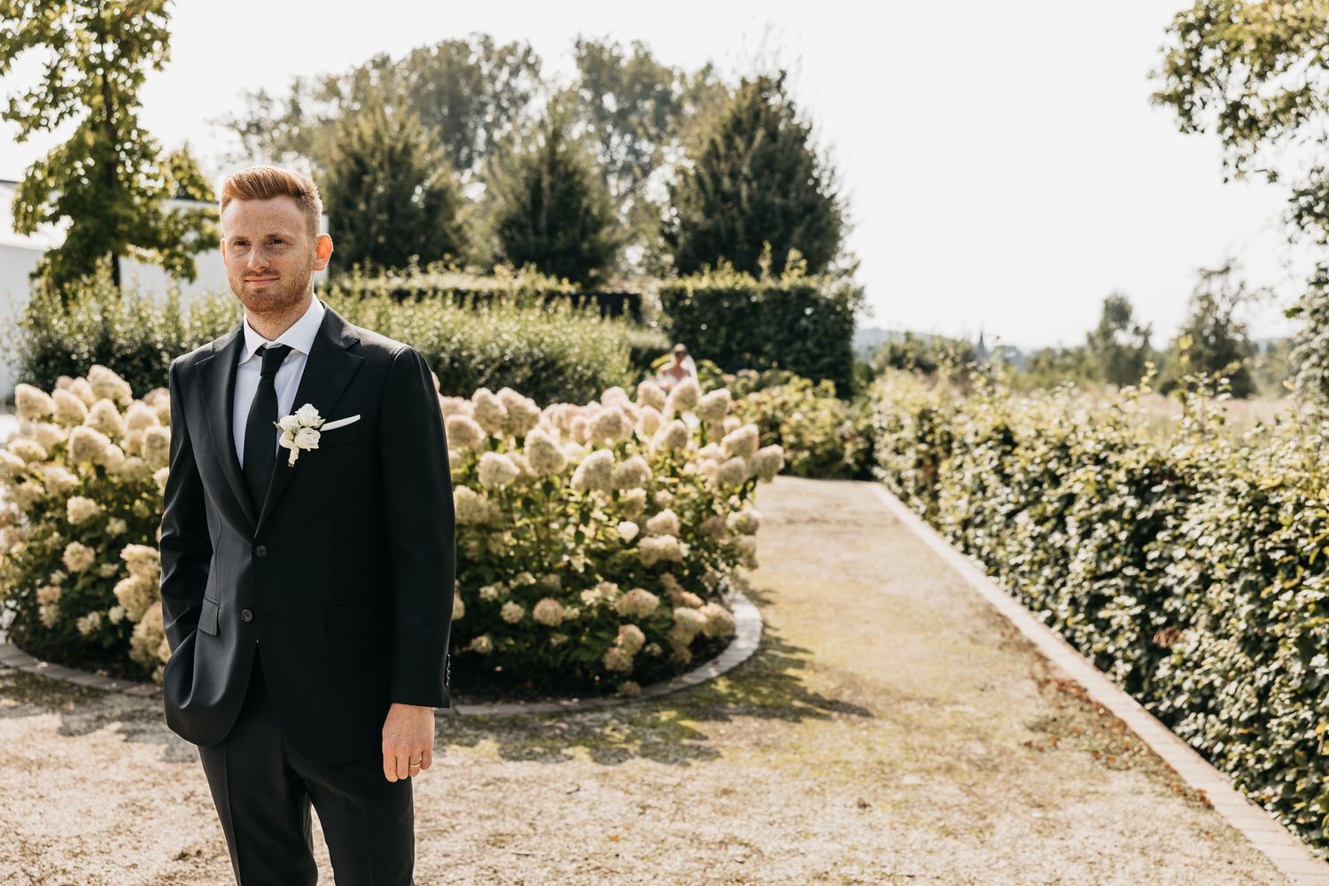 Dapper groom in black suit with white boutonniere standing in a sunlit garden path.