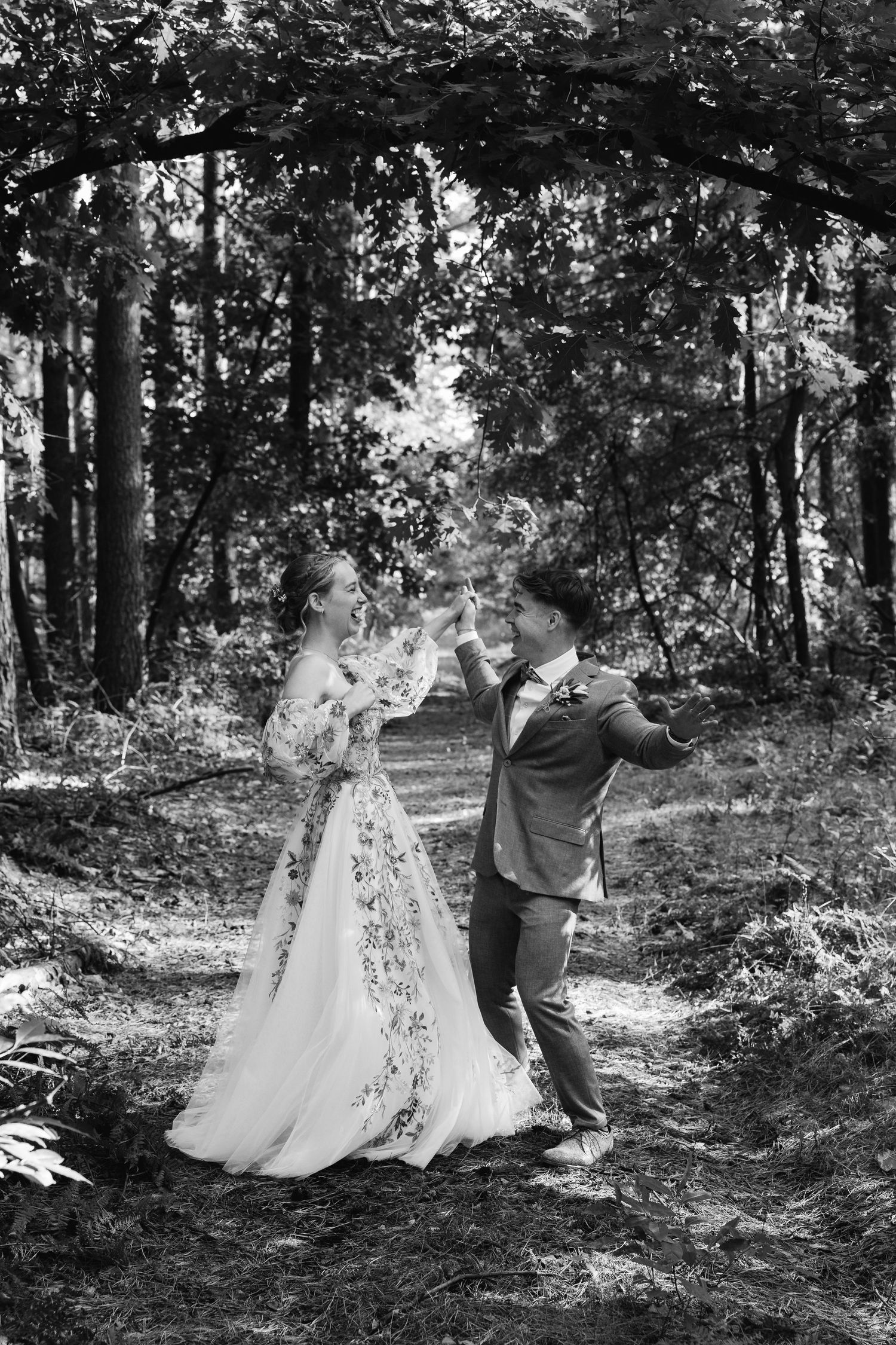 Black and white photo of bride and groom dancing on a forest path, bride in floral gown, groom in suit.