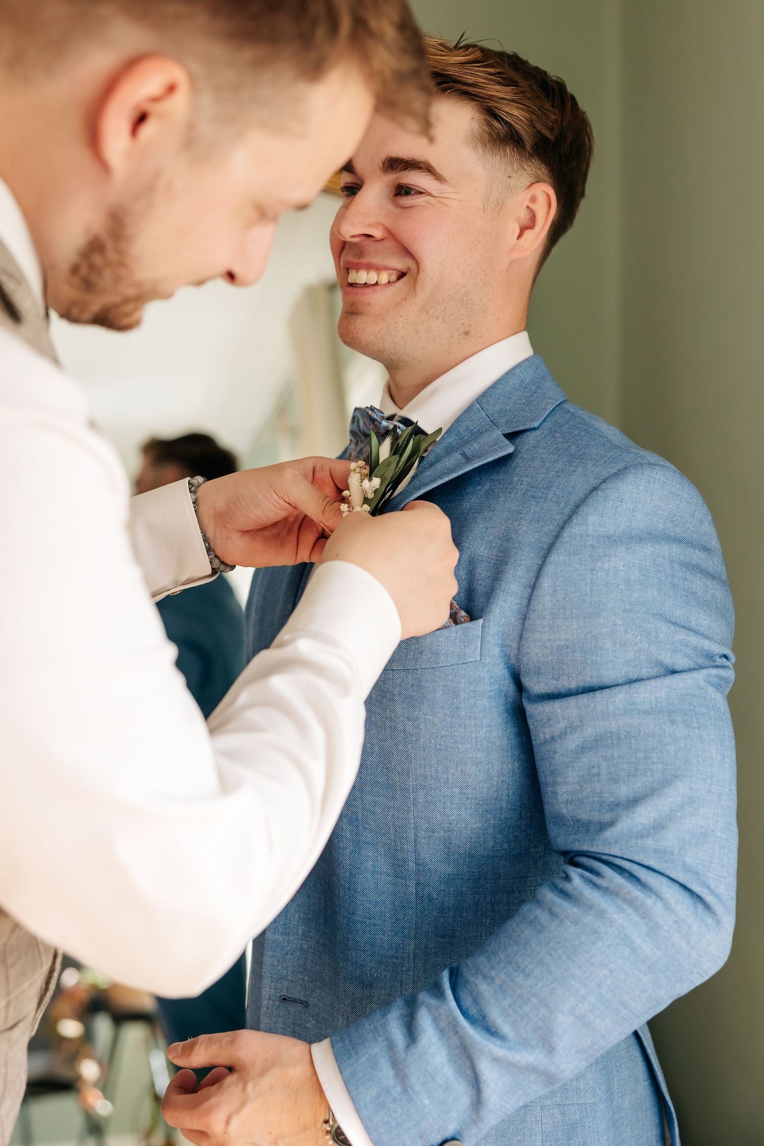 Groomsman pinning boutonniere on smiling groom in light blue wedding suit.