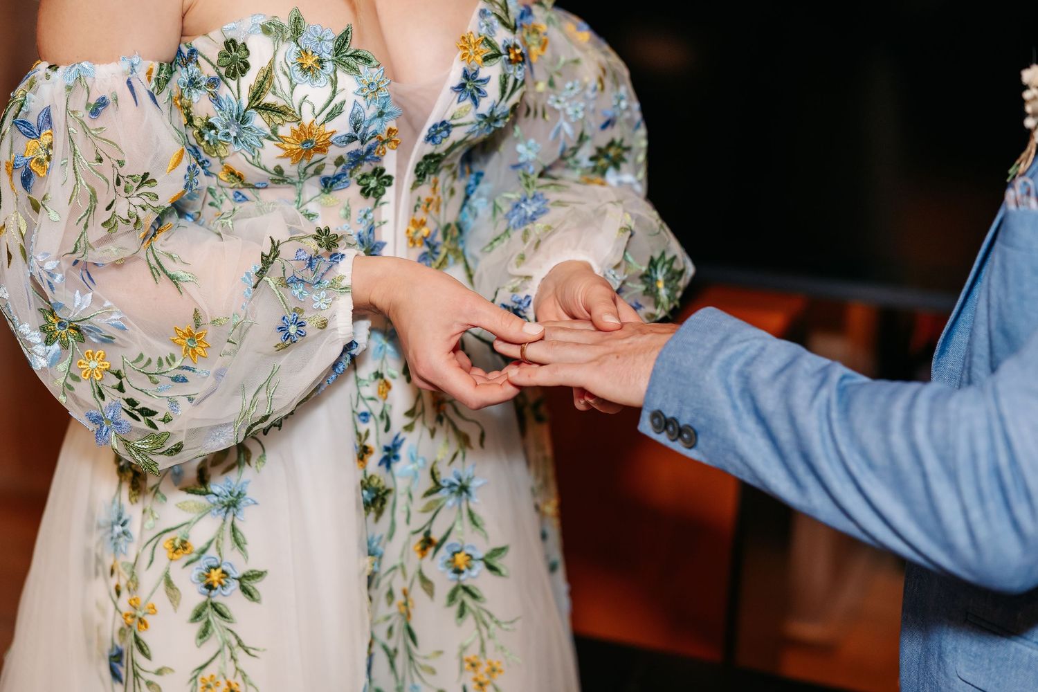 Close-up of groom sliding wedding ring onto brides finger, bride wearing floral wedding dress.