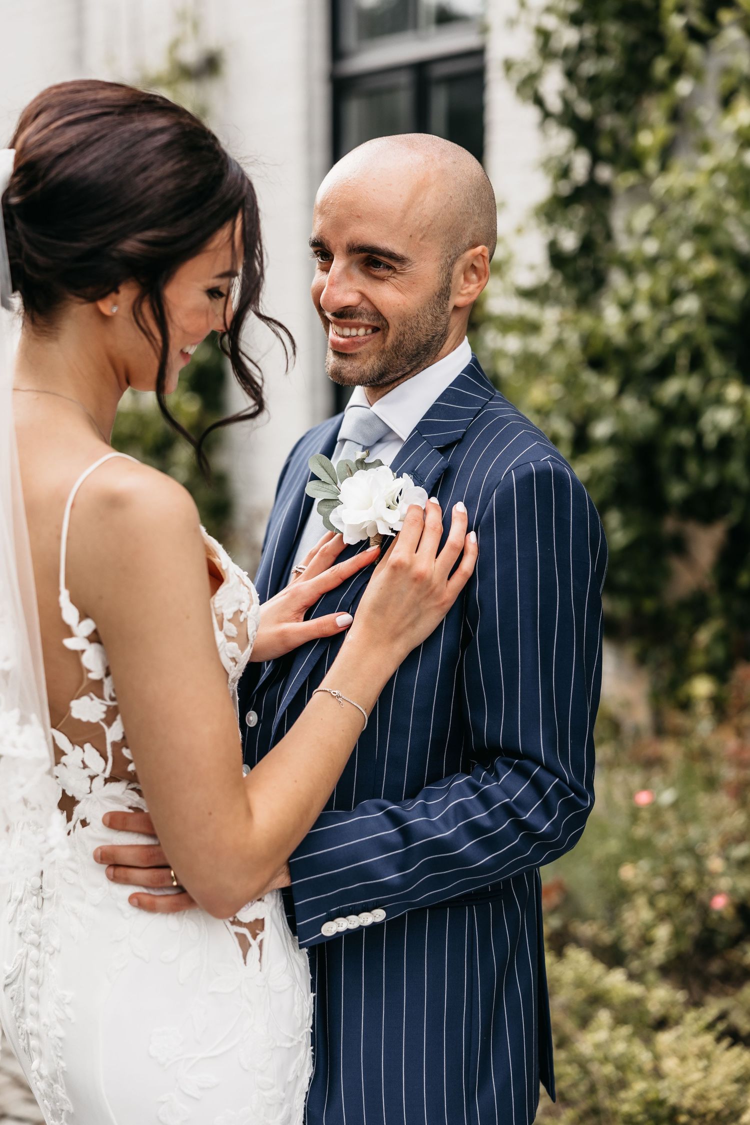 Smiling bald groom in navy pinstripe suit gazes at bride as she adjusts his boutonniere outdoors.