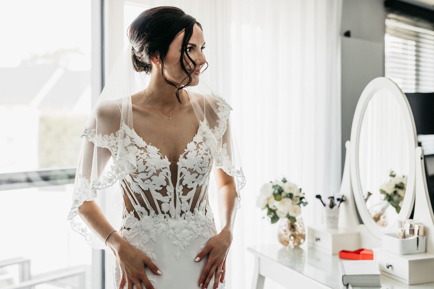Gorgeous bride in detailed lace mermaid wedding dress posing near white vanity mirror by window.
