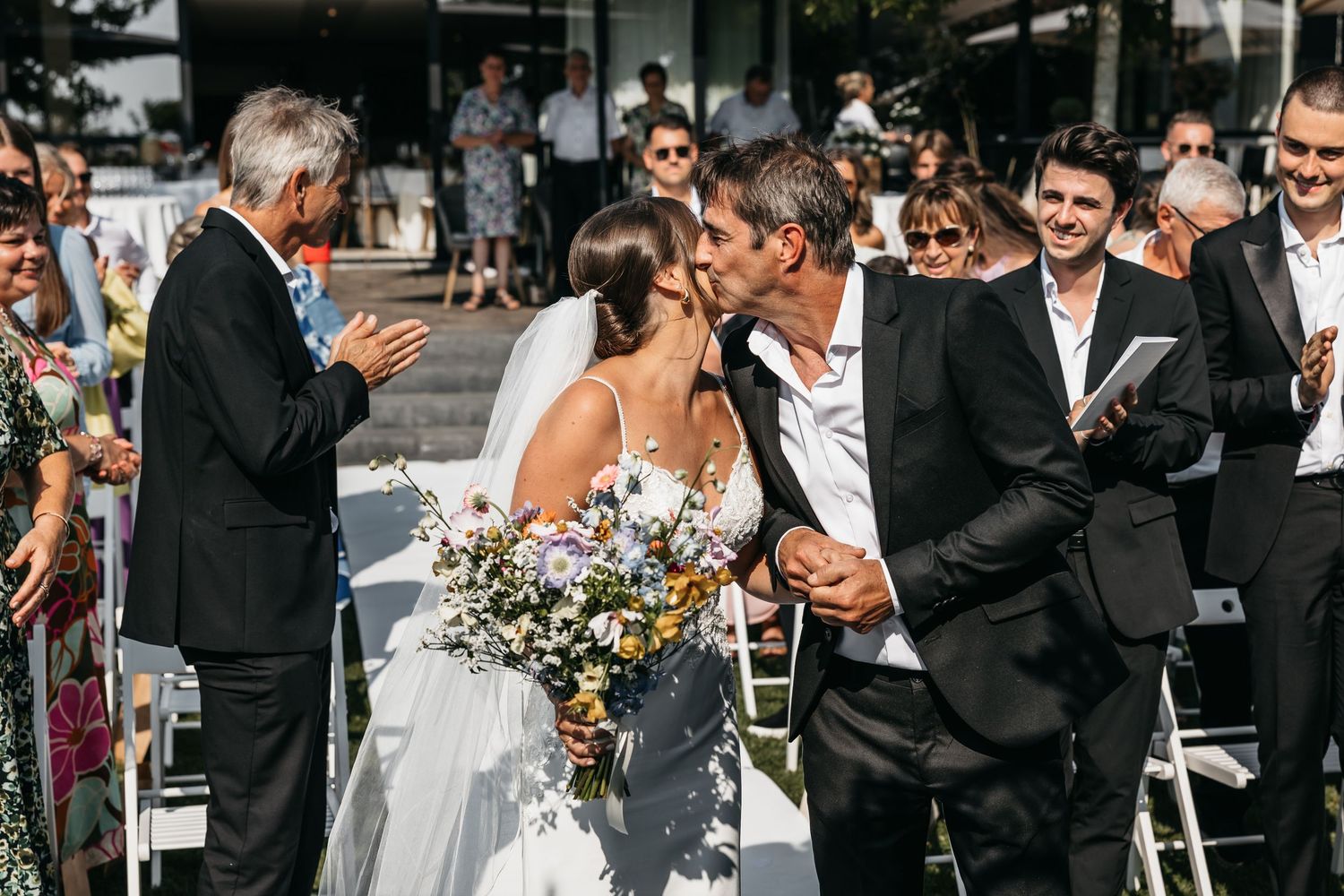 Bride and groom share a first kiss at outdoor wedding ceremony surrounded by cheering guests.