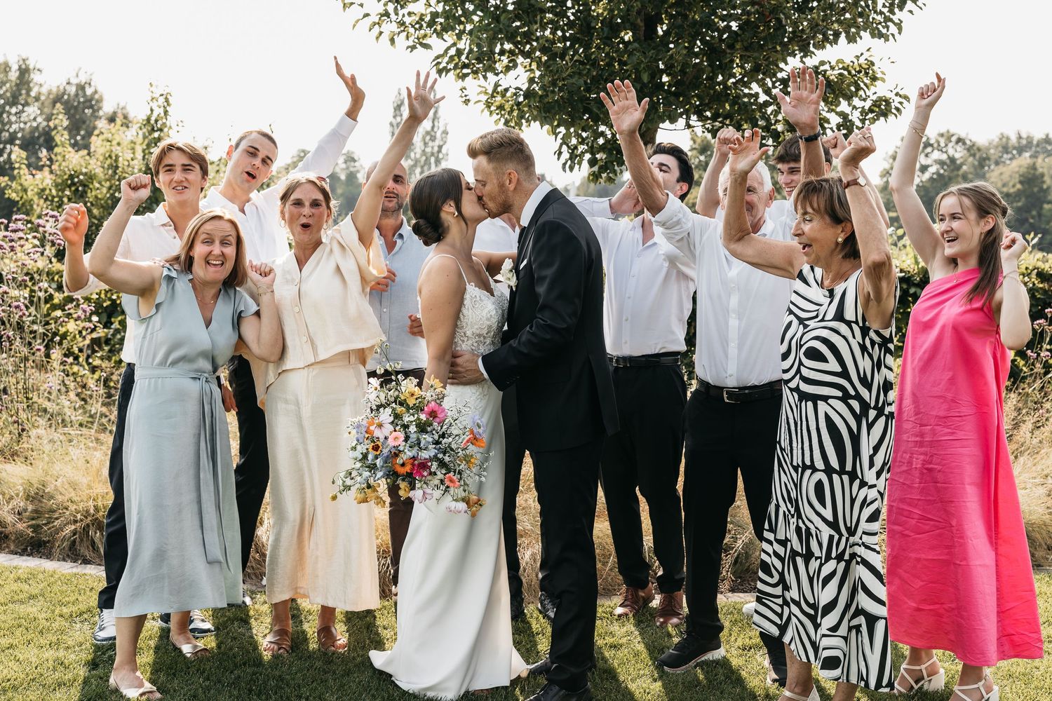 Wedding party celebrating outdoors with raised arms as bride and groom share a kiss, holding colorful bouquet.