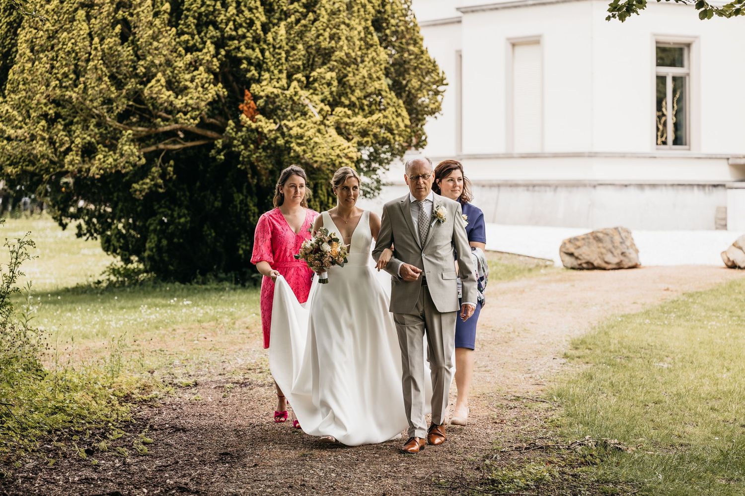 Bride in white gown walks with two escorts along garden path toward outdoor wedding ceremony venue.