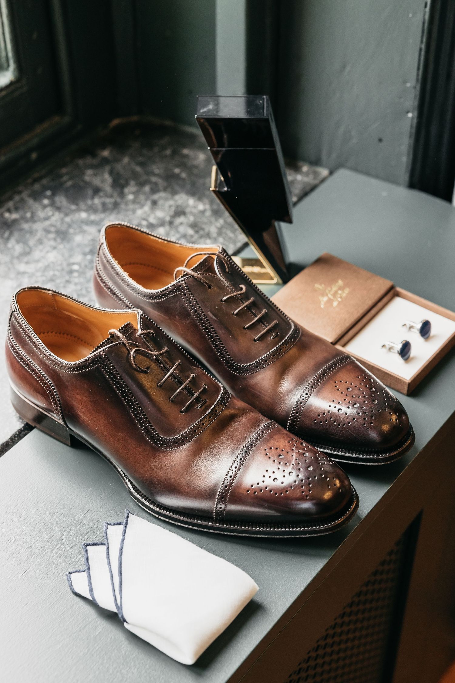 Brown leather brogue Oxford dress shoes with cufflinks and pocket square on a dark surface.