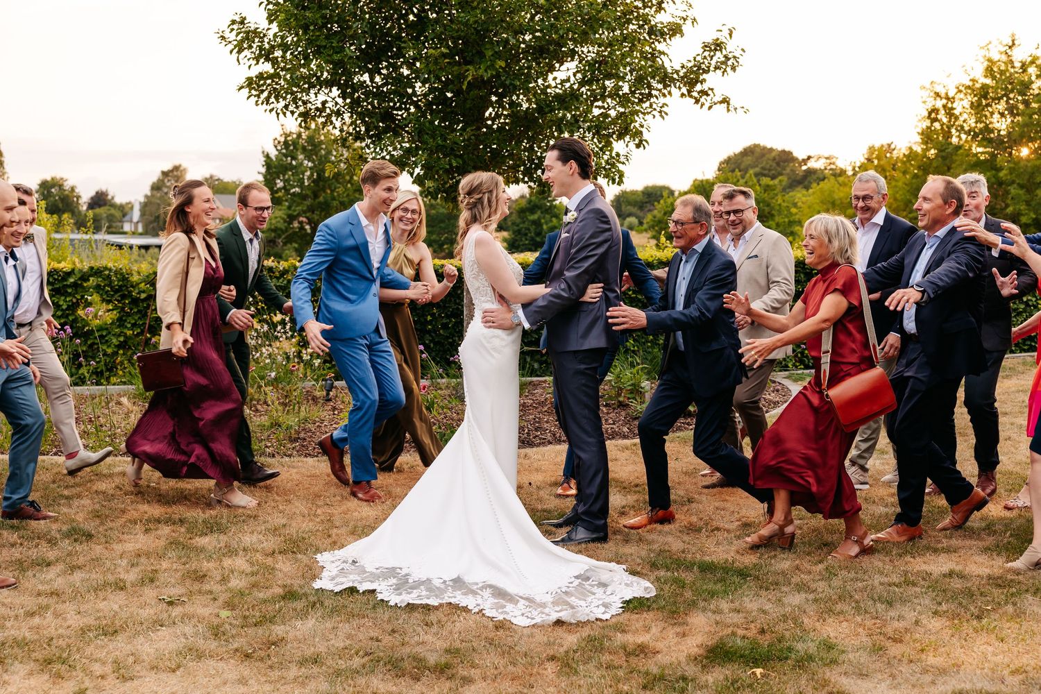 Bride and groom celebrate joyfully with wedding guests dancing together in a garden at sunset.