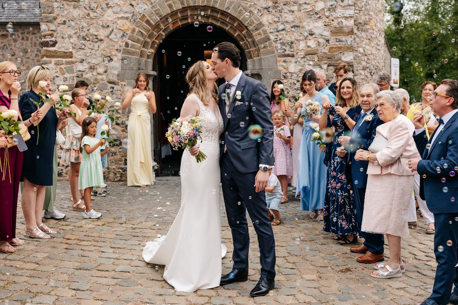 Bride and groom share a kiss outside stone church surrounded by guests blowing bubbles.