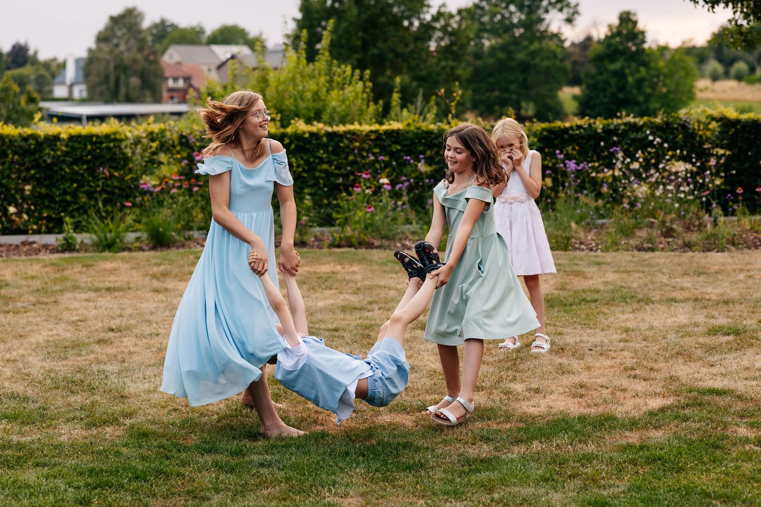 Women in light blue dresses playfully swing a child by the arms in a lush garden at a wedding.