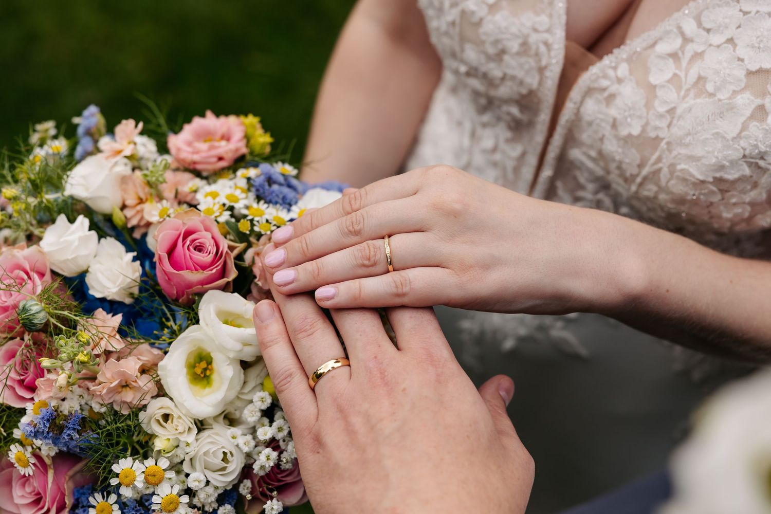 Newlyweds holding hands displaying matching gold wedding rings over colorful bridal bouquet.