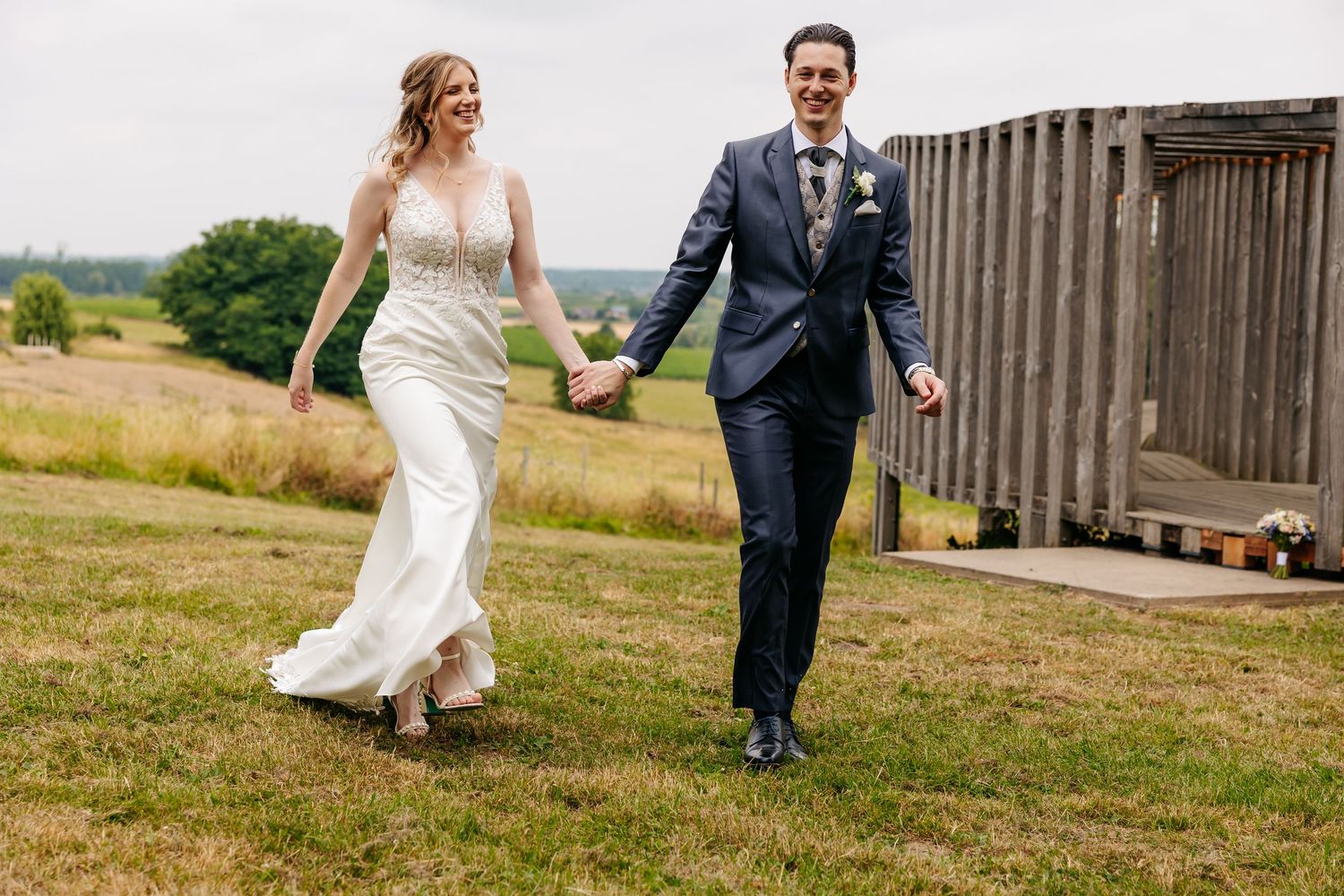 Joyful bride and groom holding hands and walking together outdoors near a rustic wooden structure on their wedding day.