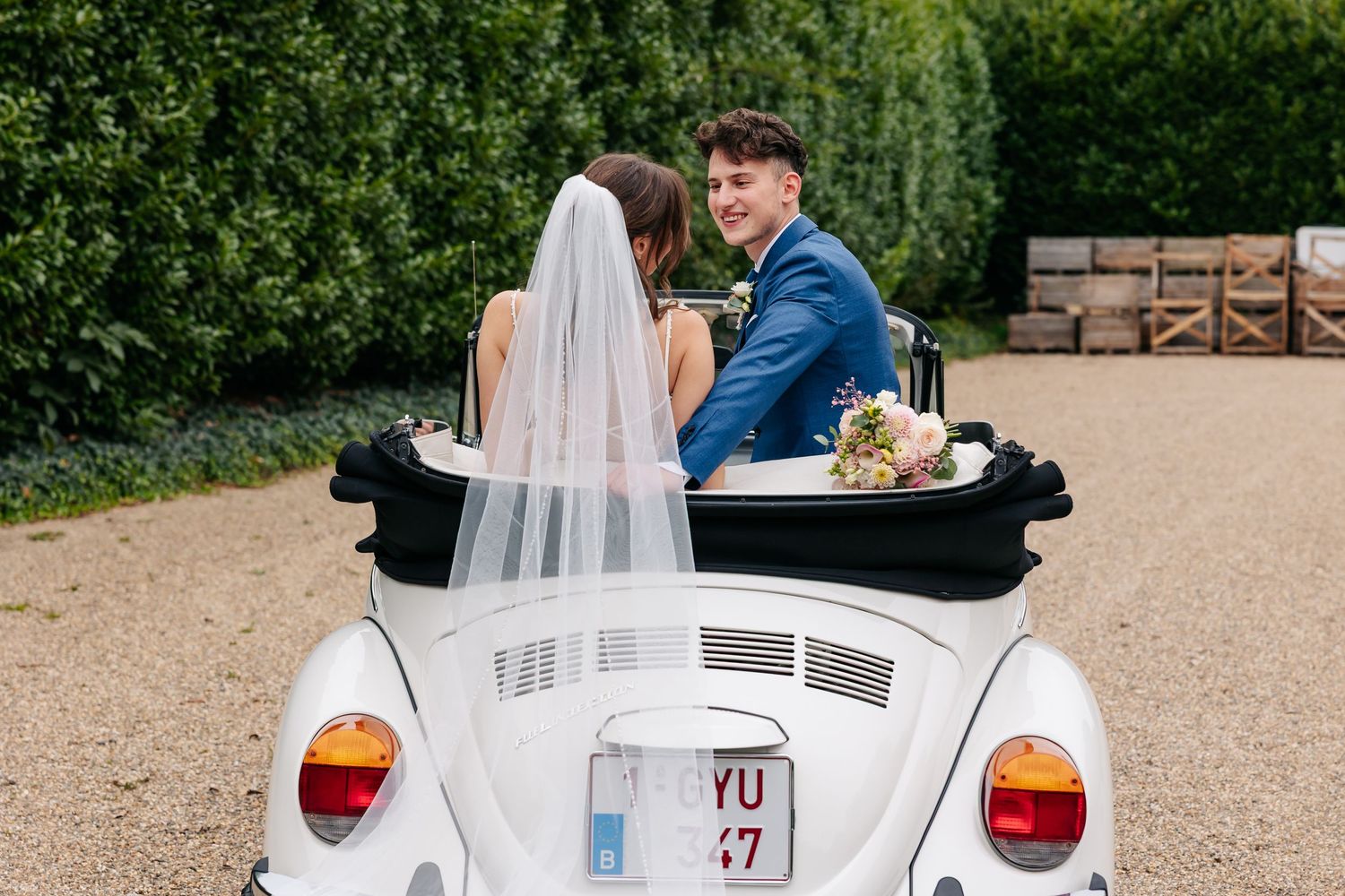 Smiling bride and groom pose in white vintage VW Beetle convertible with bouquet on wedding day.