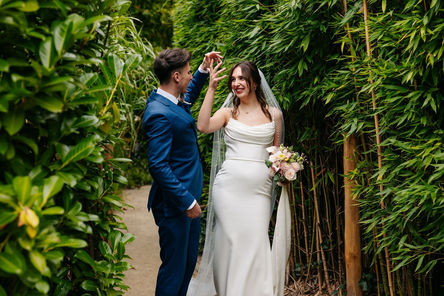 Groom in blue suit twirling bride in white gown among tall green bamboo plants outdoors.
