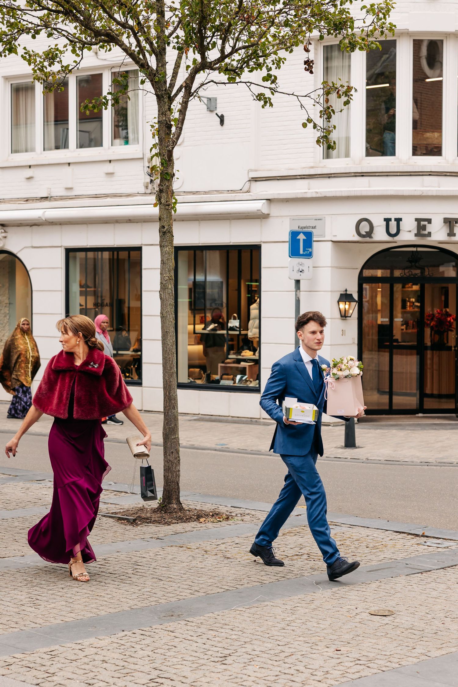 Man in blue suit carrying flowers and woman in burgundy velvet gown walking along a European city street.