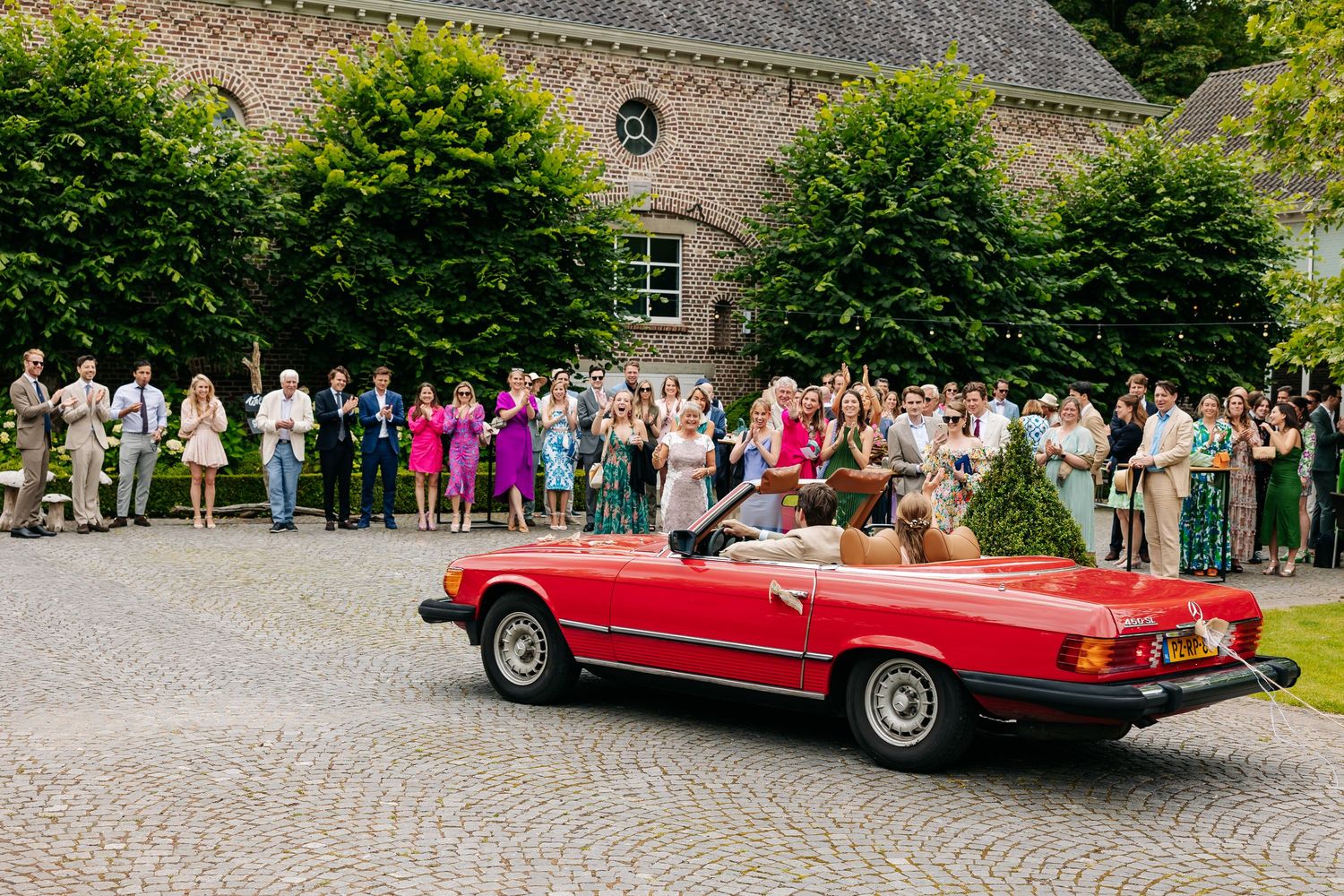 Red convertible classic car driving through cobblestone courtyard with wedding guests cheering alongside.