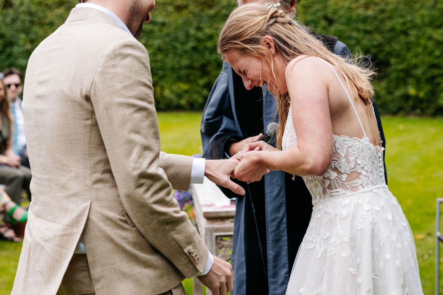 Bride in lace wedding gown placing a ring on the groom's finger during an outdoor wedding ceremony.