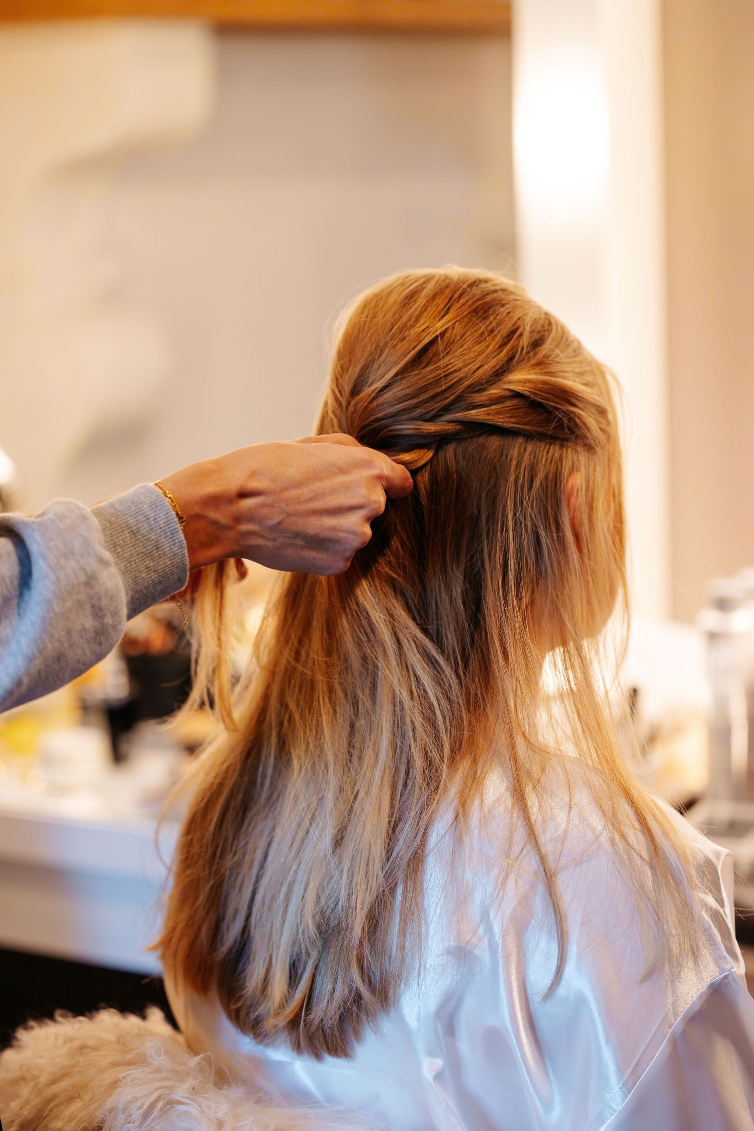 A hairstylist styling a blonde woman's hair into an elegant half-updo in a bright salon mirror.