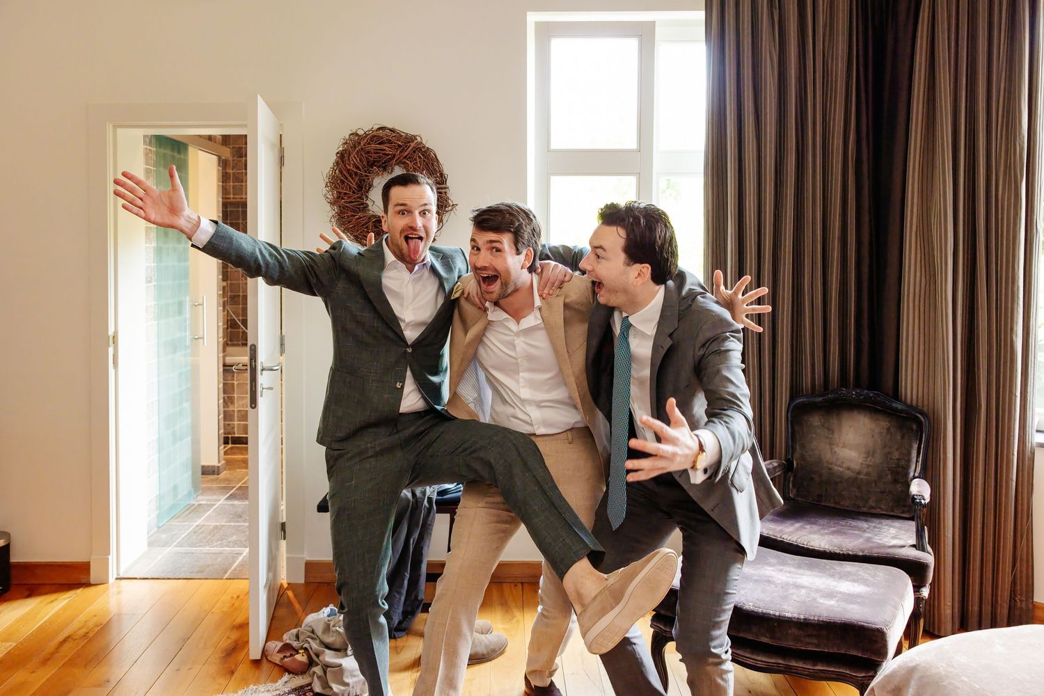 Three groomsmen in suits celebrating joyfully in hotel room before wedding.