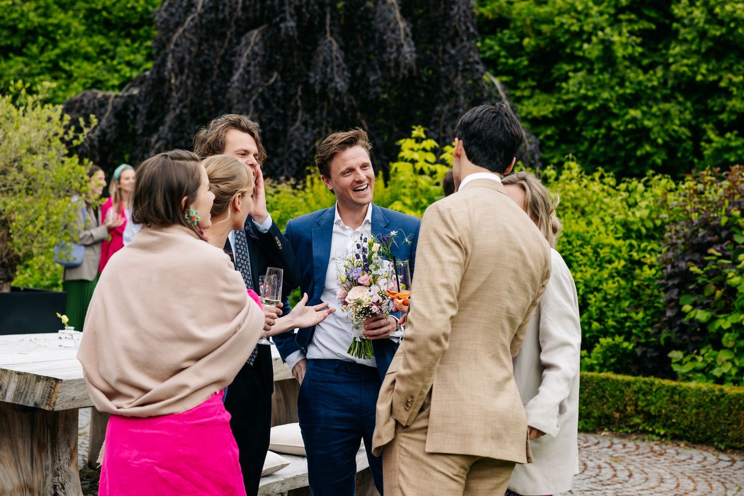 Guests laughing with man holding bouquet at elegant outdoor garden wedding cocktail hour.