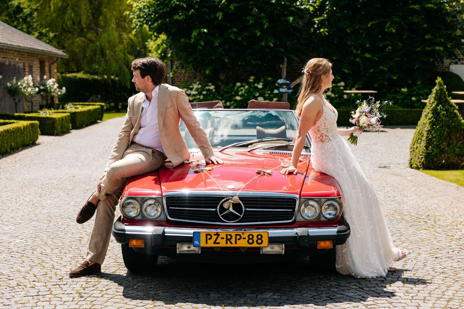 Bride and groom posing with a vintage red Mercedes-Benz convertible on a cobblestone driveway.