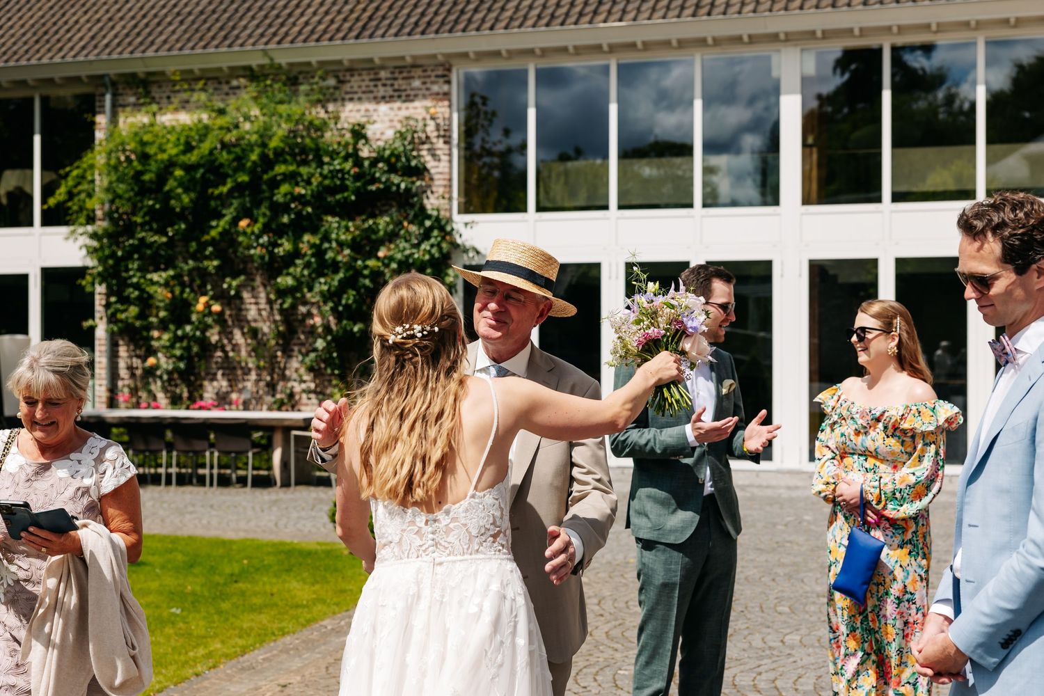 Bride in white lace gown greeting a male guest in a straw hat outside a bright glass-windowed wedding venue.