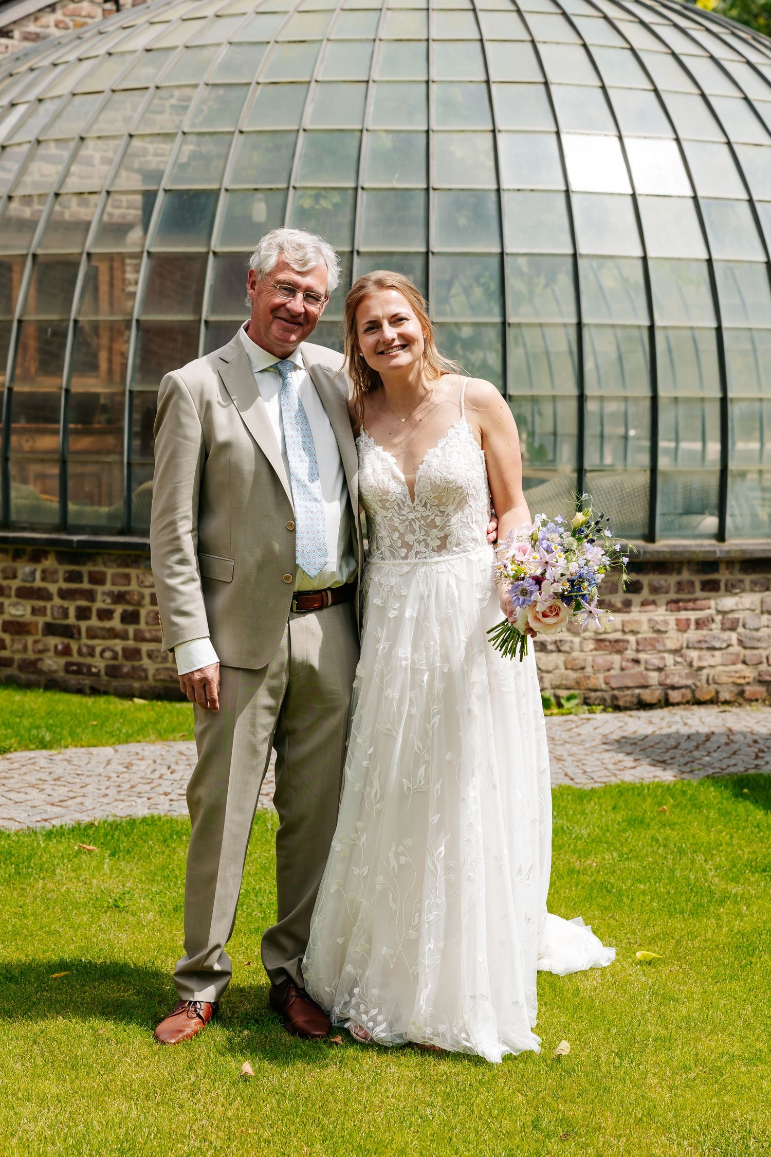 Bride holding bouquet poses with older man in tan suit before glass dome greenhouse.