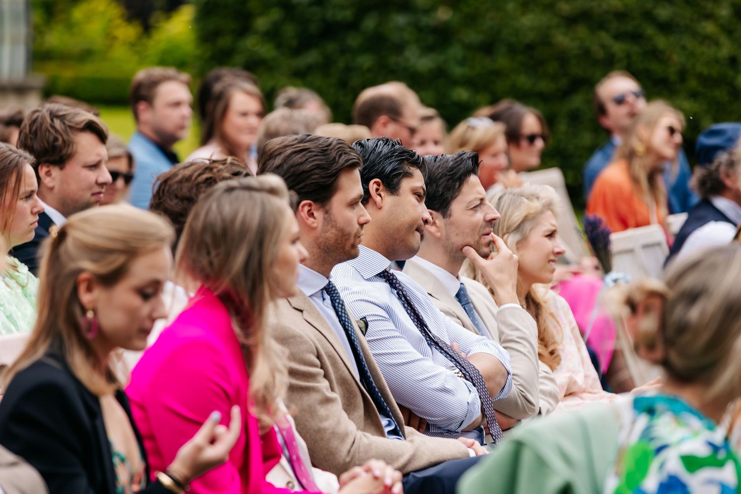 Elegantly dressed wedding guests seated outdoors enjoying a sunny garden wedding ceremony.