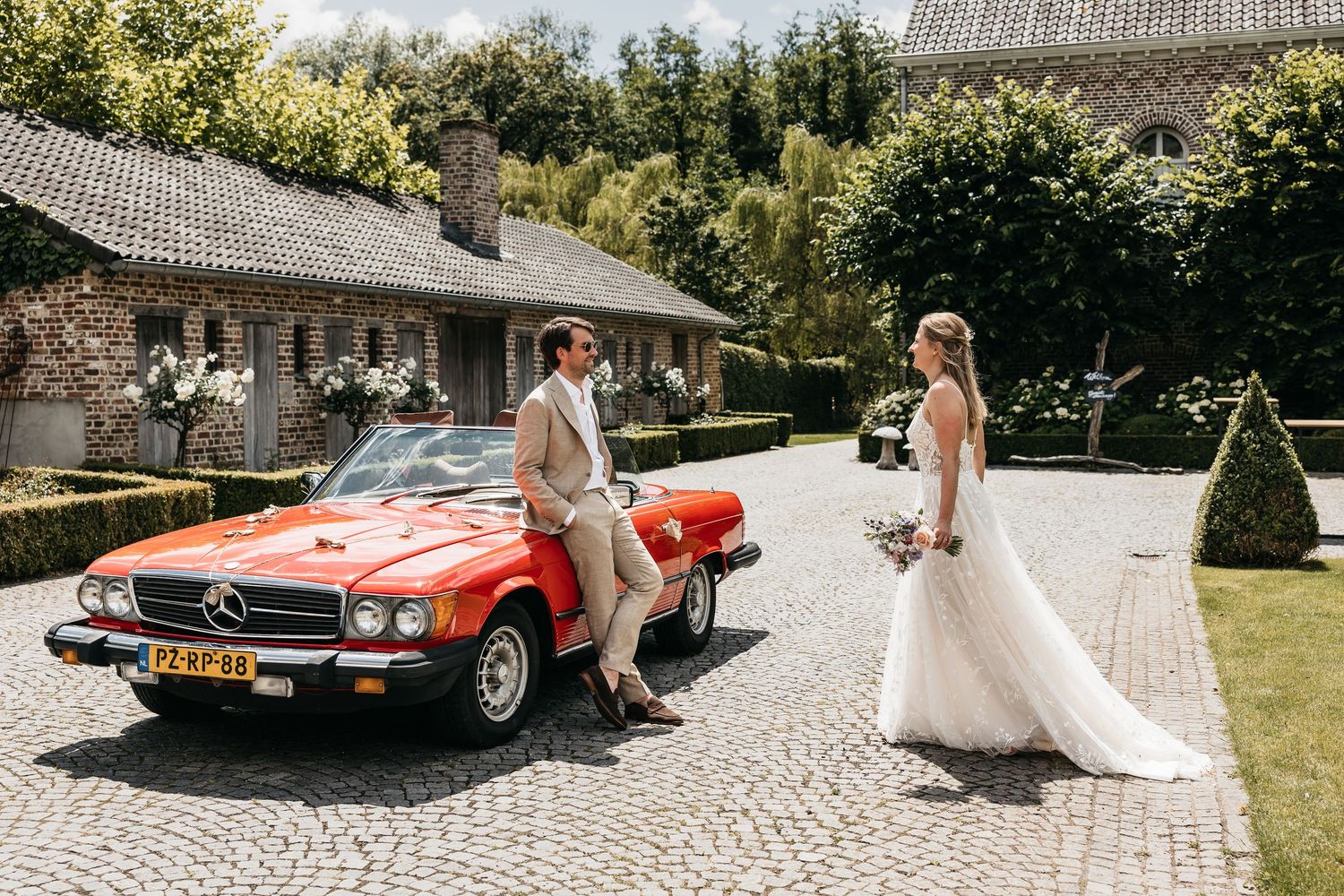 Bride and groom standing apart by a vintage red Mercedes convertible on sunny wedding day.
