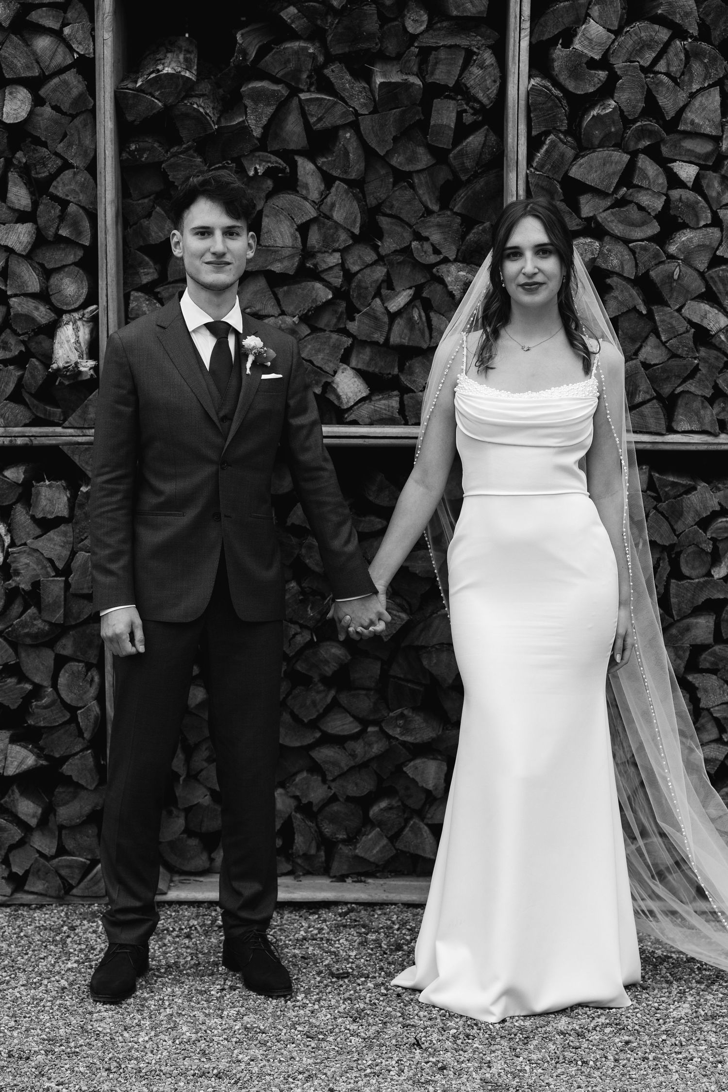Black and white wedding portrait of bride and groom holding hands before a rustic stacked wood wall.