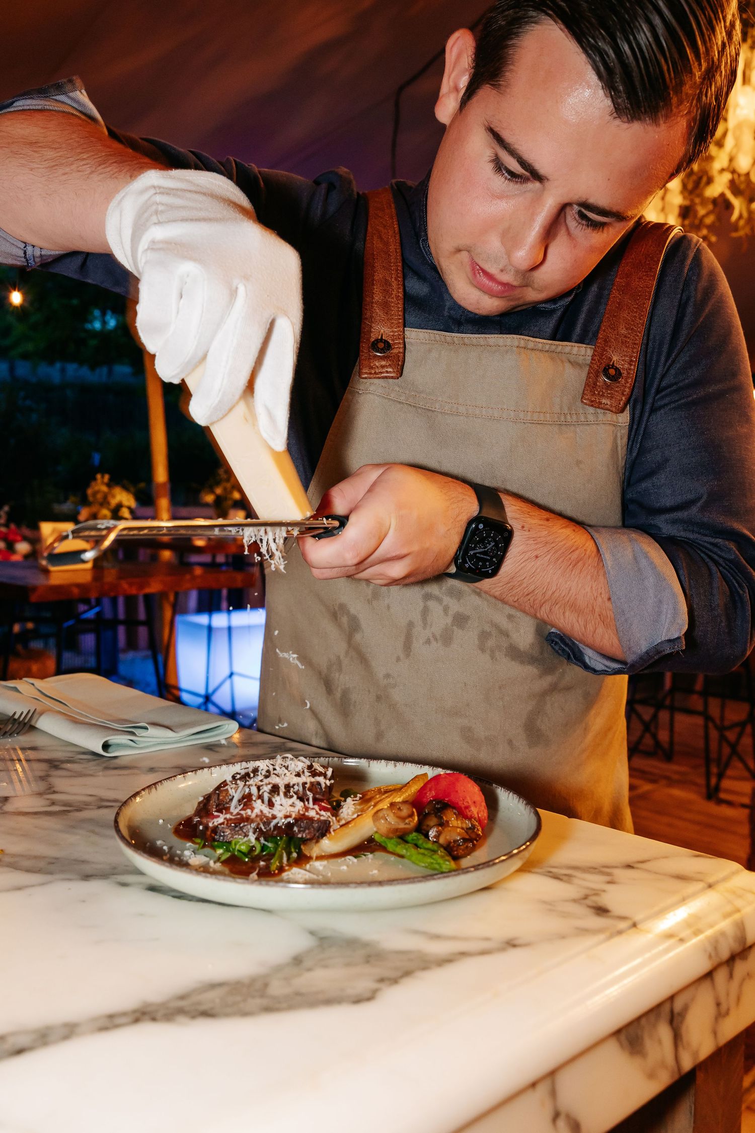 Chef in apron carefully grating fresh parmesan cheese over a gourmet plated dish at marble counter.