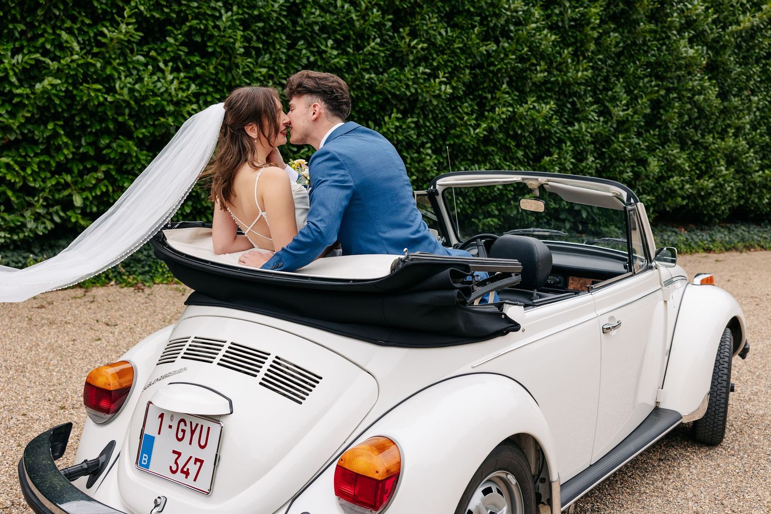 Bride and groom share a kiss in a white convertible VW Beetle with veil flowing in the breeze.
