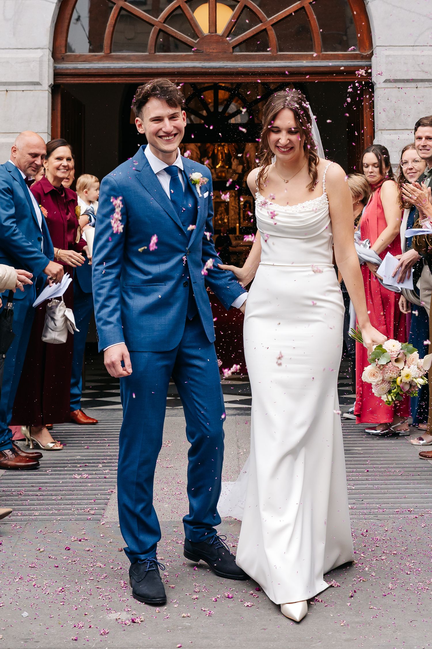 Newlyweds walk out of church smiling as guests throw pink confetti, bride in white gown, groom in blue suit.