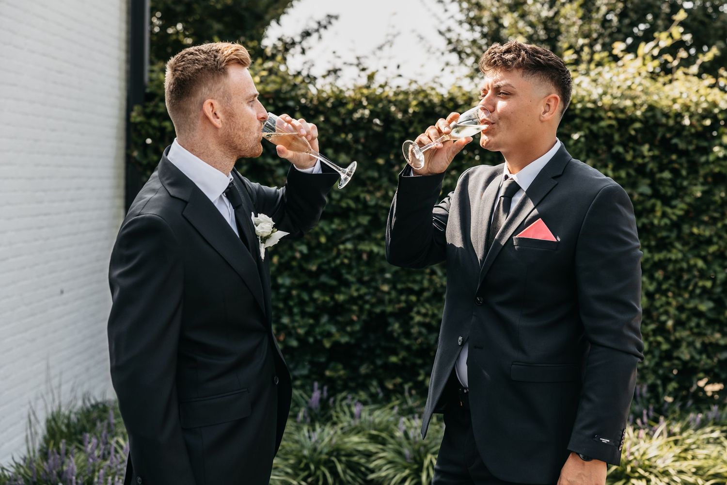 Two groomsmen in black suits drink champagne together in a garden before a wedding.