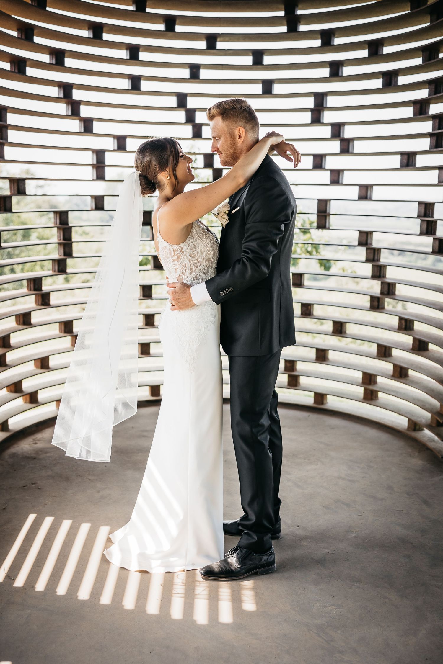 Bride in a lace wedding gown and groom in a black suit embrace inside a curved wooden chapel interior.