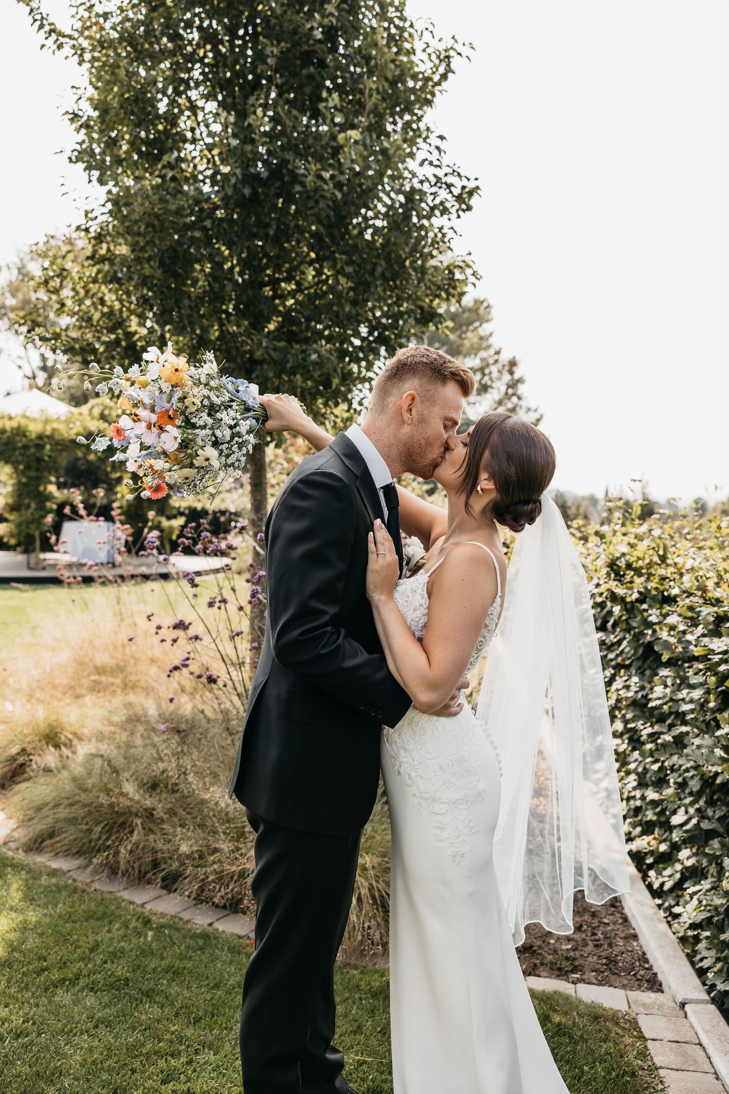 Bride and groom sharing a romantic kiss in a garden, bride holding a colorful wildflower bouquet.
