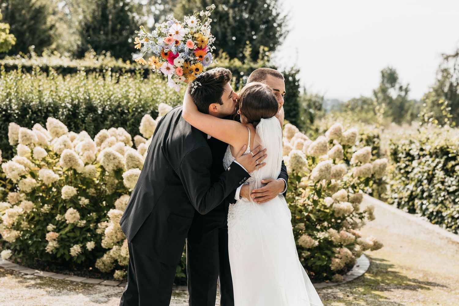Groom in black suit embraces bride holding colorful bouquet among white hydrangeas at outdoor wedding.