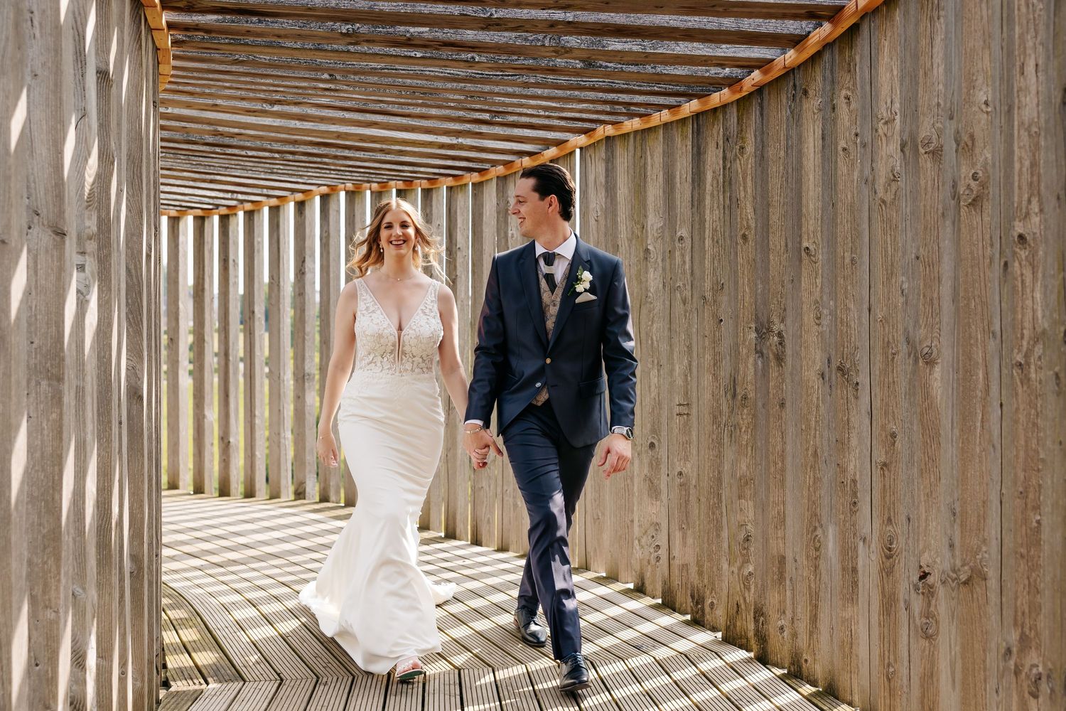 Bride in fitted lace wedding gown and groom in navy suit walking hand in hand through wooden pergola.