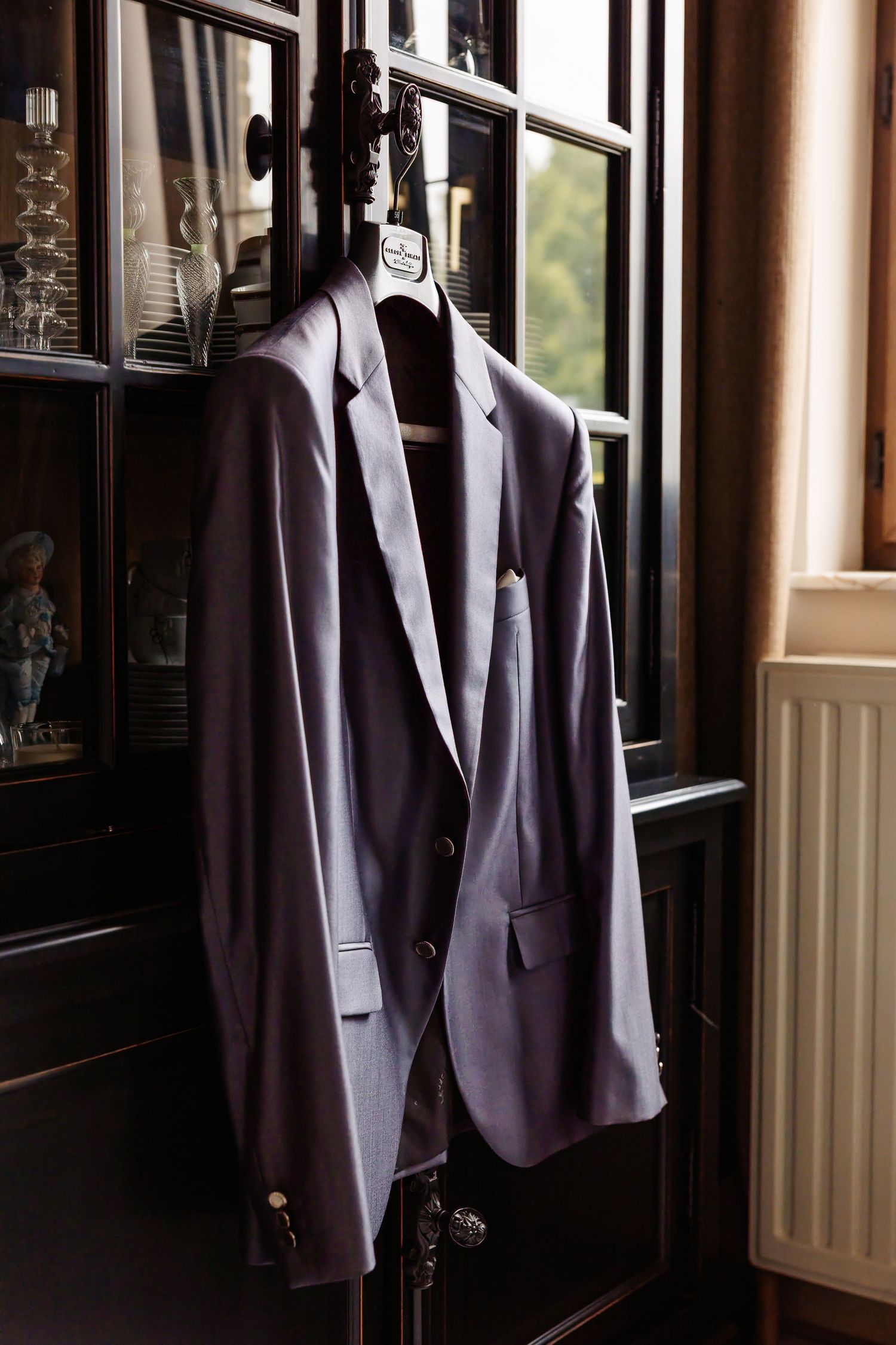 Elegant gray grooms suit jacket hanging on decorative wooden hanger beside dark wood cabinet.