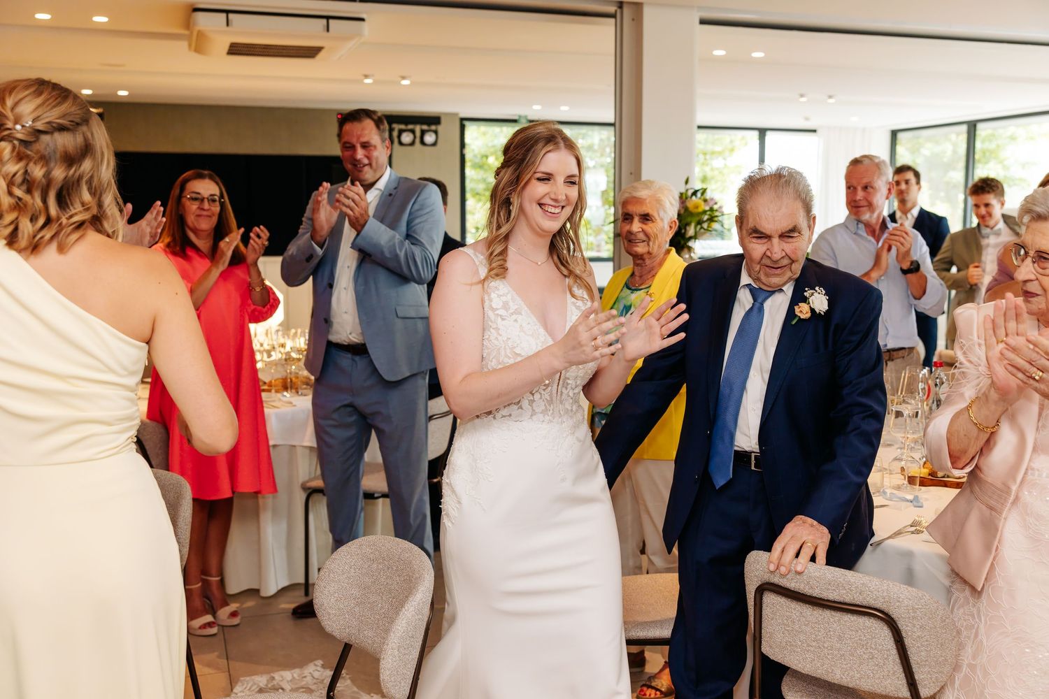 Bride in white gown enters wedding reception with older gentleman as guests clap and celebrate.
