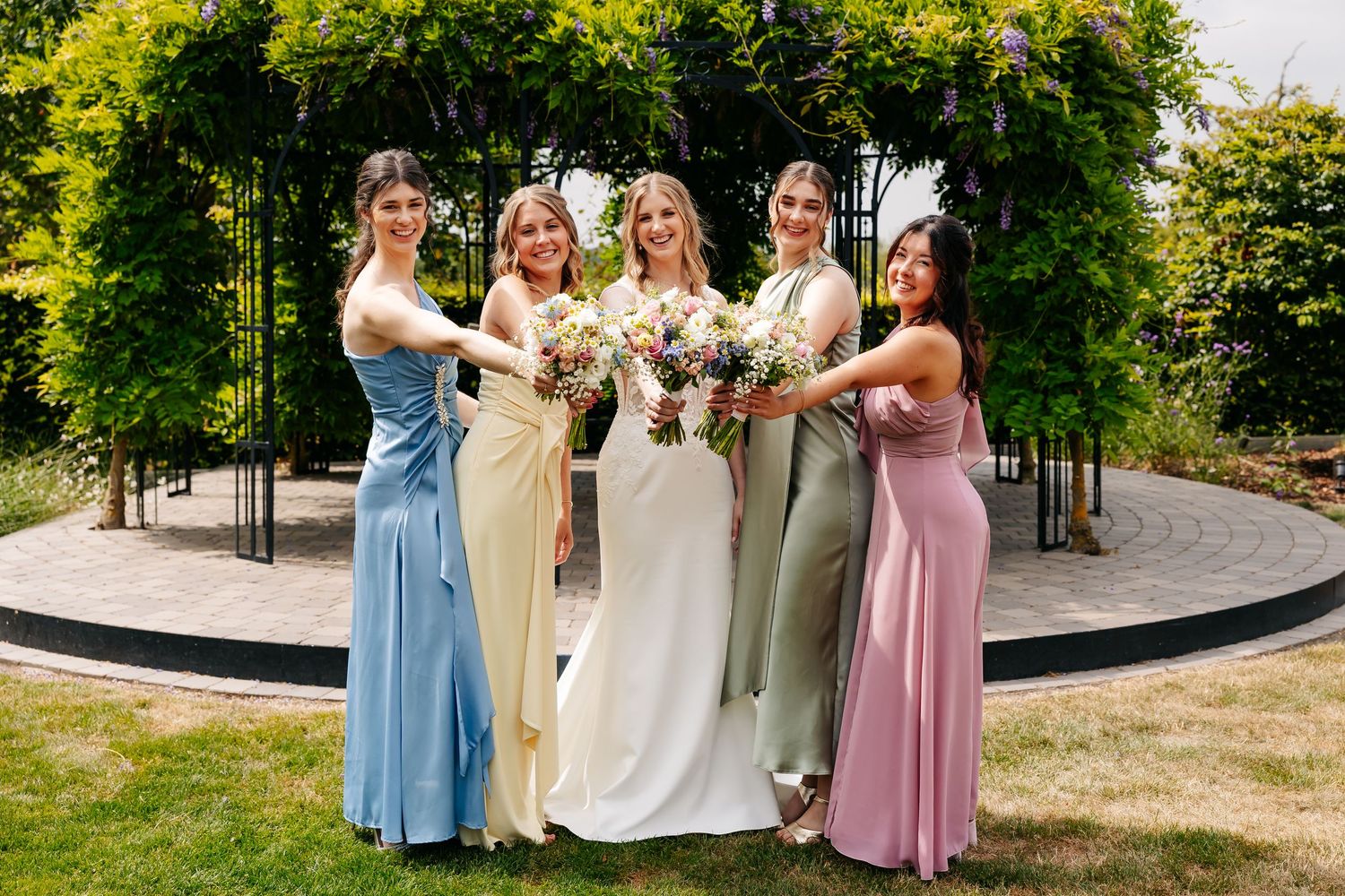 Bride and four bridesmaids in colorful pastel dresses smile and hold bouquets together in a garden pergola.