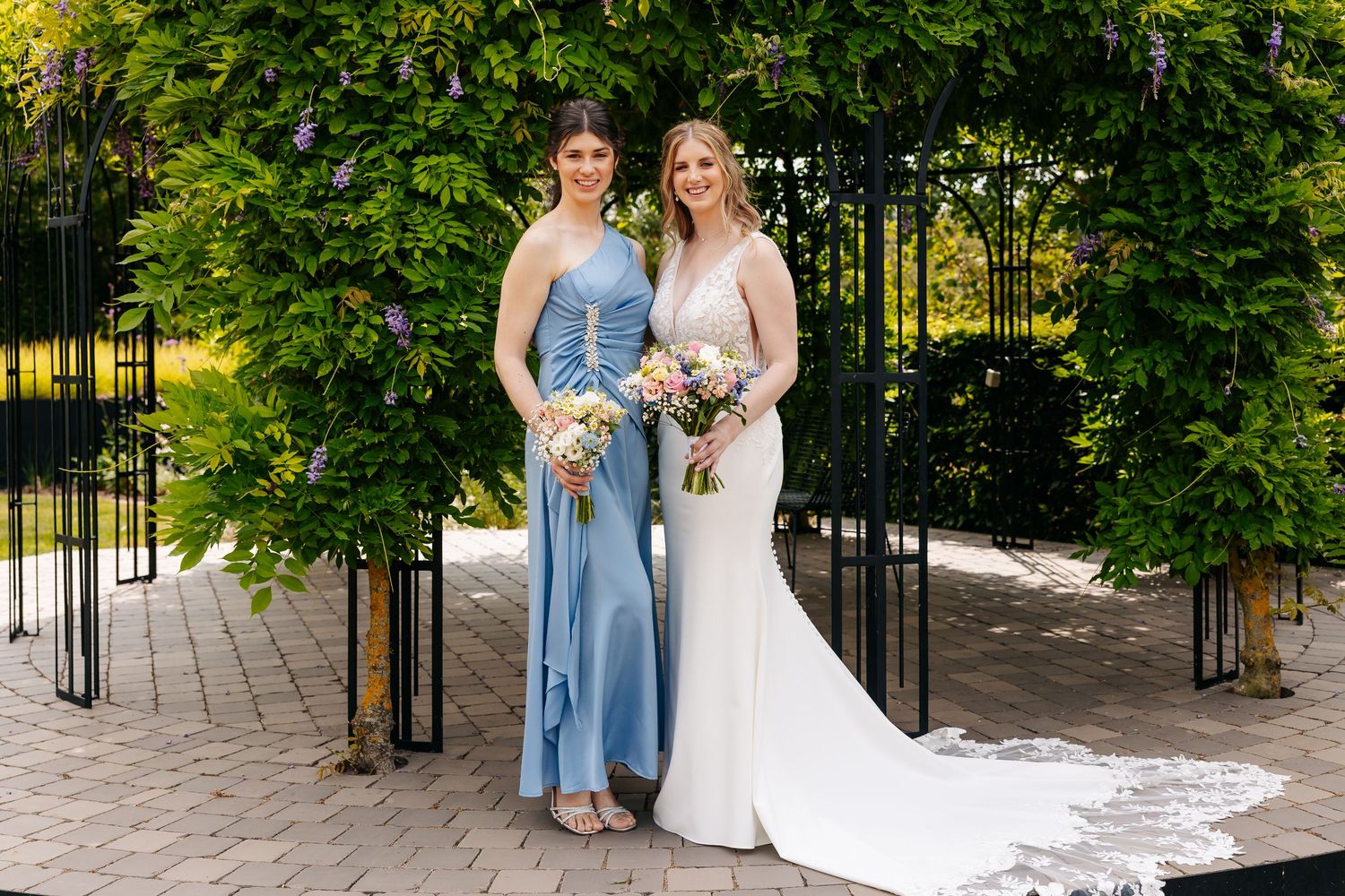 Bride in fitted white gown and bridesmaid in blue dress posing together under green garden archway.