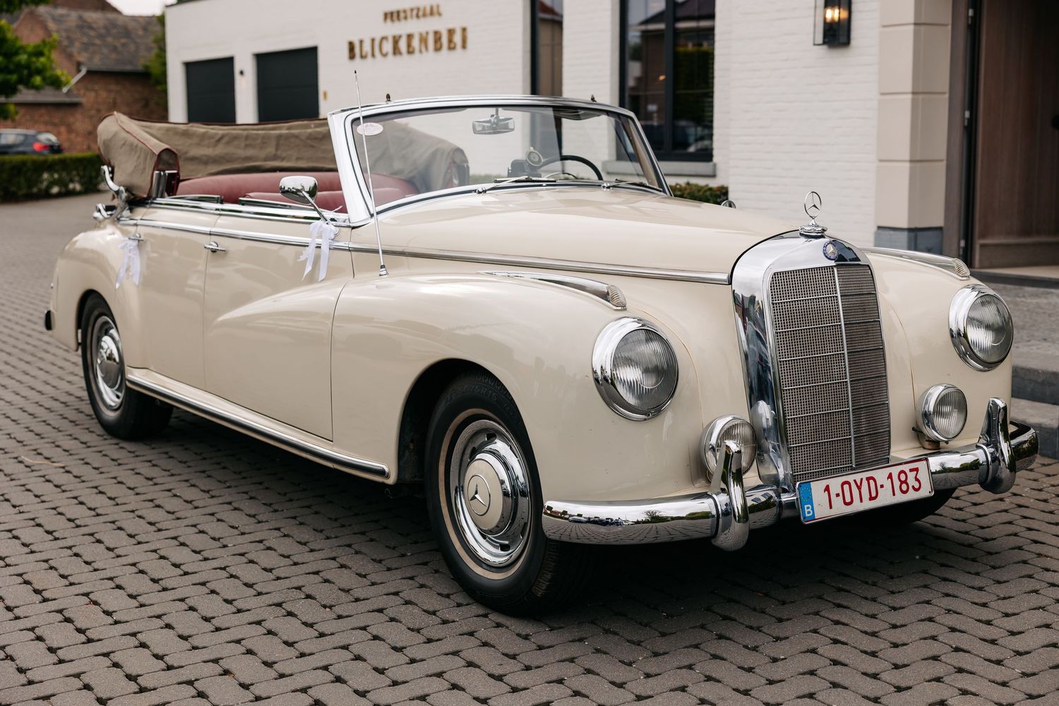 Vintage cream Mercedes-Benz convertible wedding car with red interior parked outside Blickenberg venue on cobblestone.