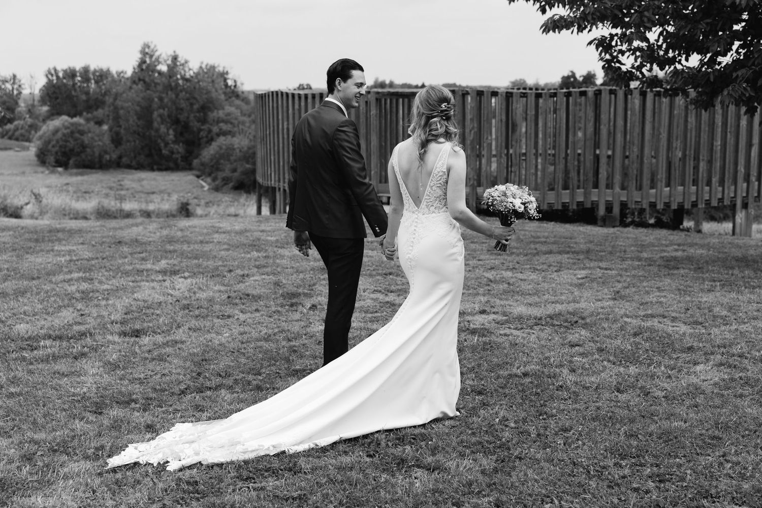Black and white photo of bride in backless gown and groom walking through a green meadow together.