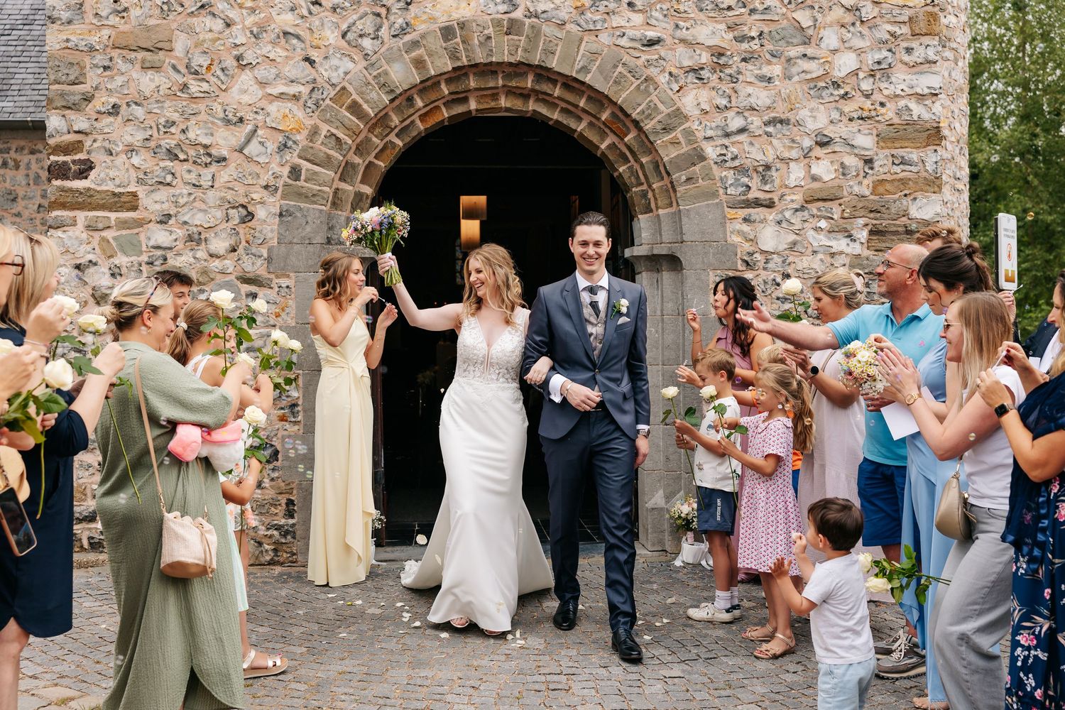 Newlyweds exit stone church arch as guests celebrate with flowers and confetti in sunny outdoor wedding.