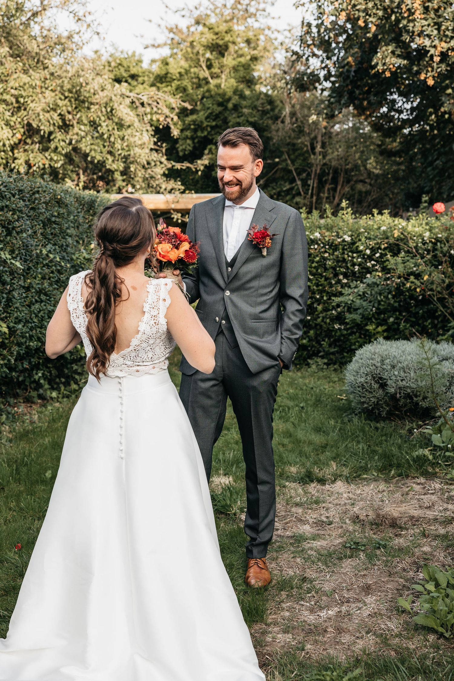 Smiling groom in gray suit facing bride holding vibrant red bouquet in sunlit garden.