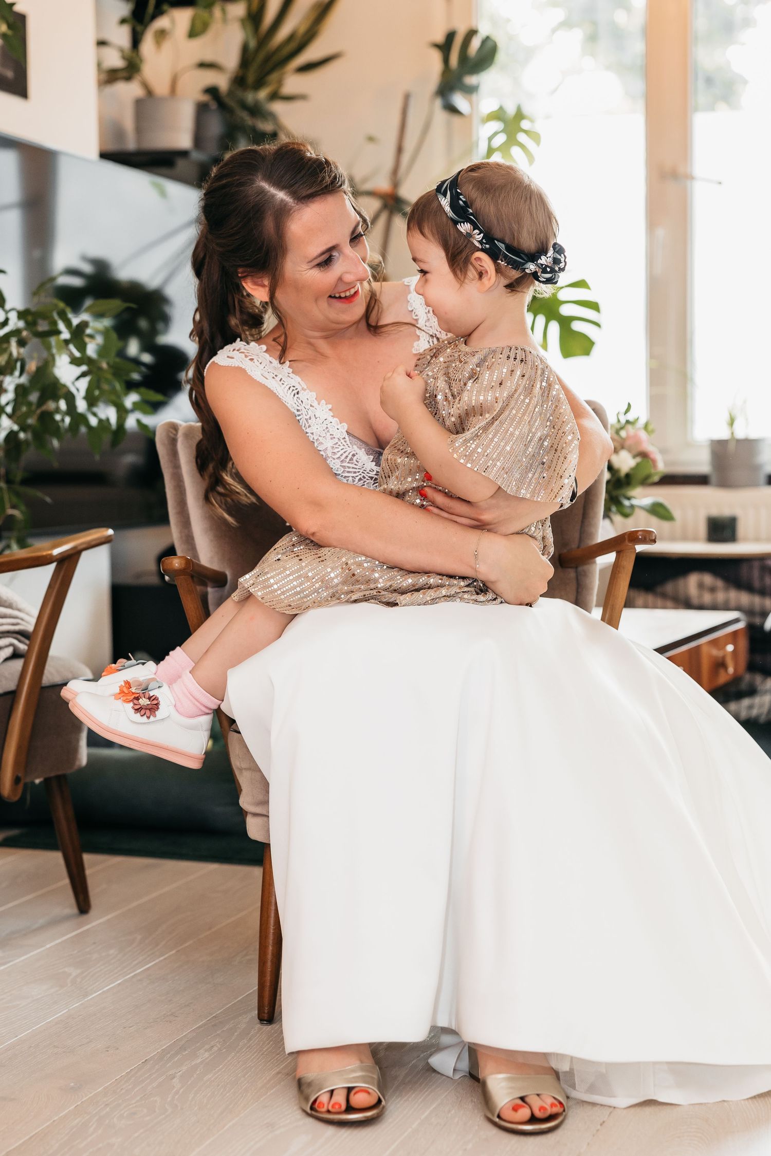 Smiling bride in white wedding dress holding a young girl in a gold sequin dress on her lap.