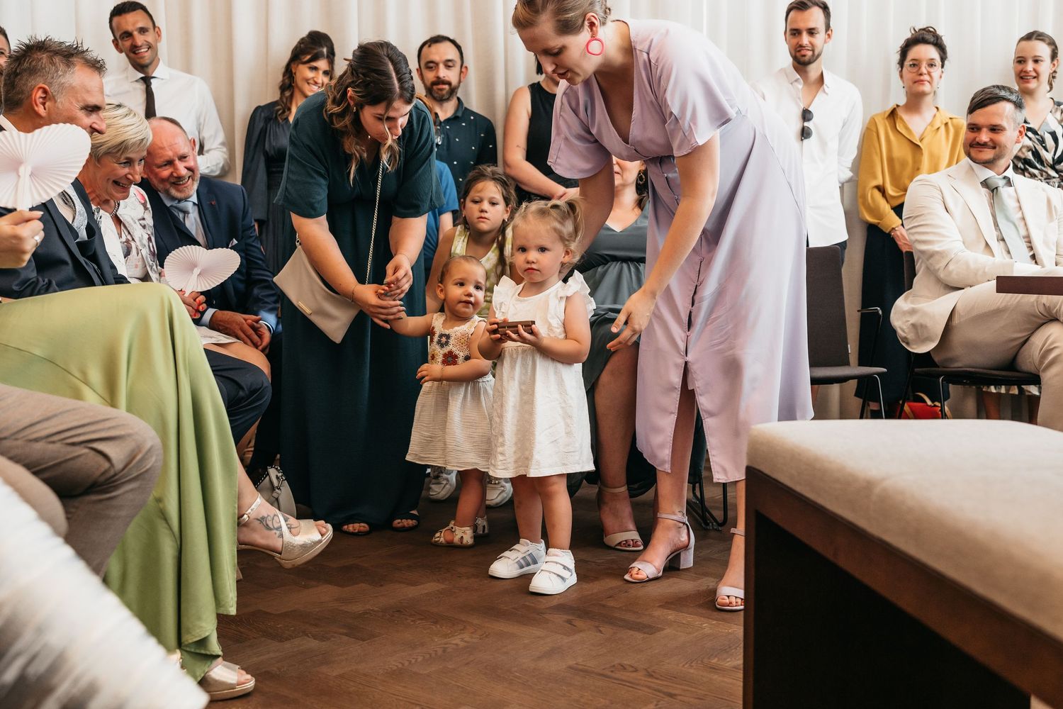 Young toddler in white dress stands with wedding guests during a ceremony while adults gather around.