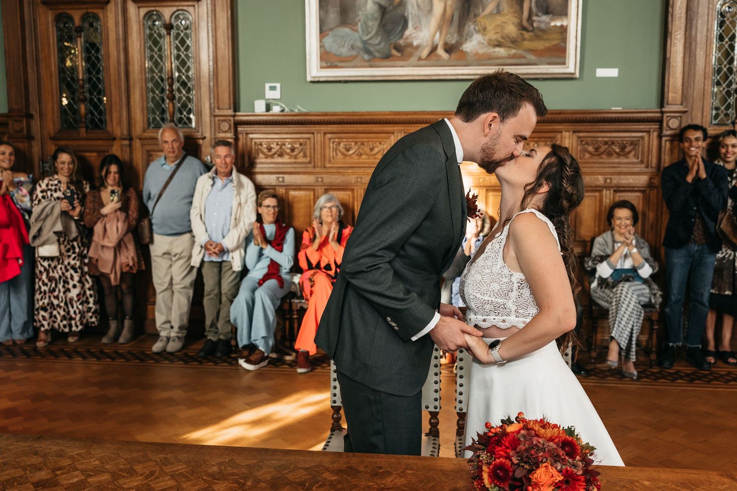 Newlyweds share their first kiss surrounded by applauding guests in a grand historic ceremony room.