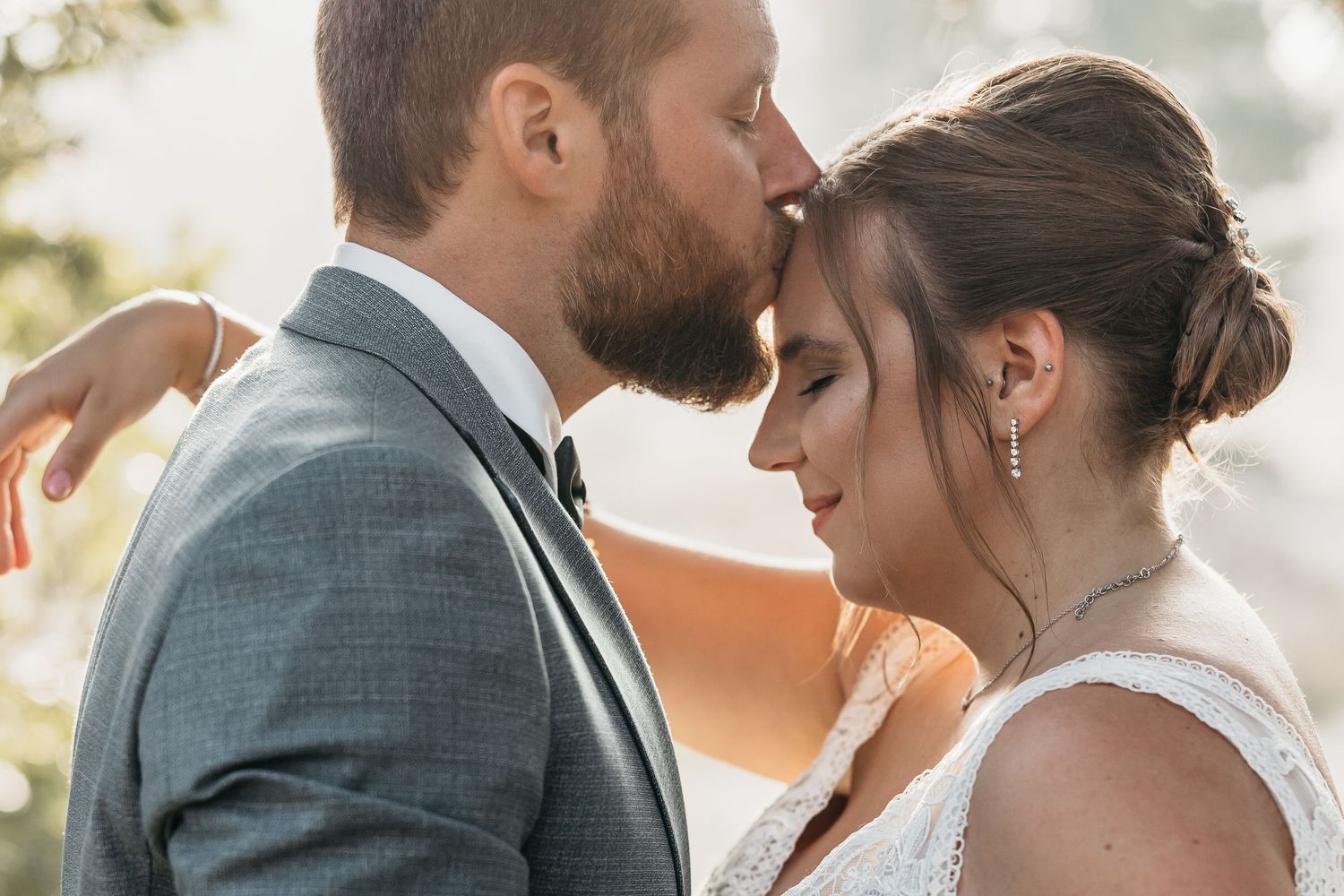 Bearded groom in gray suit kisses bride's forehead in a close romantic wedding portrait outdoors.